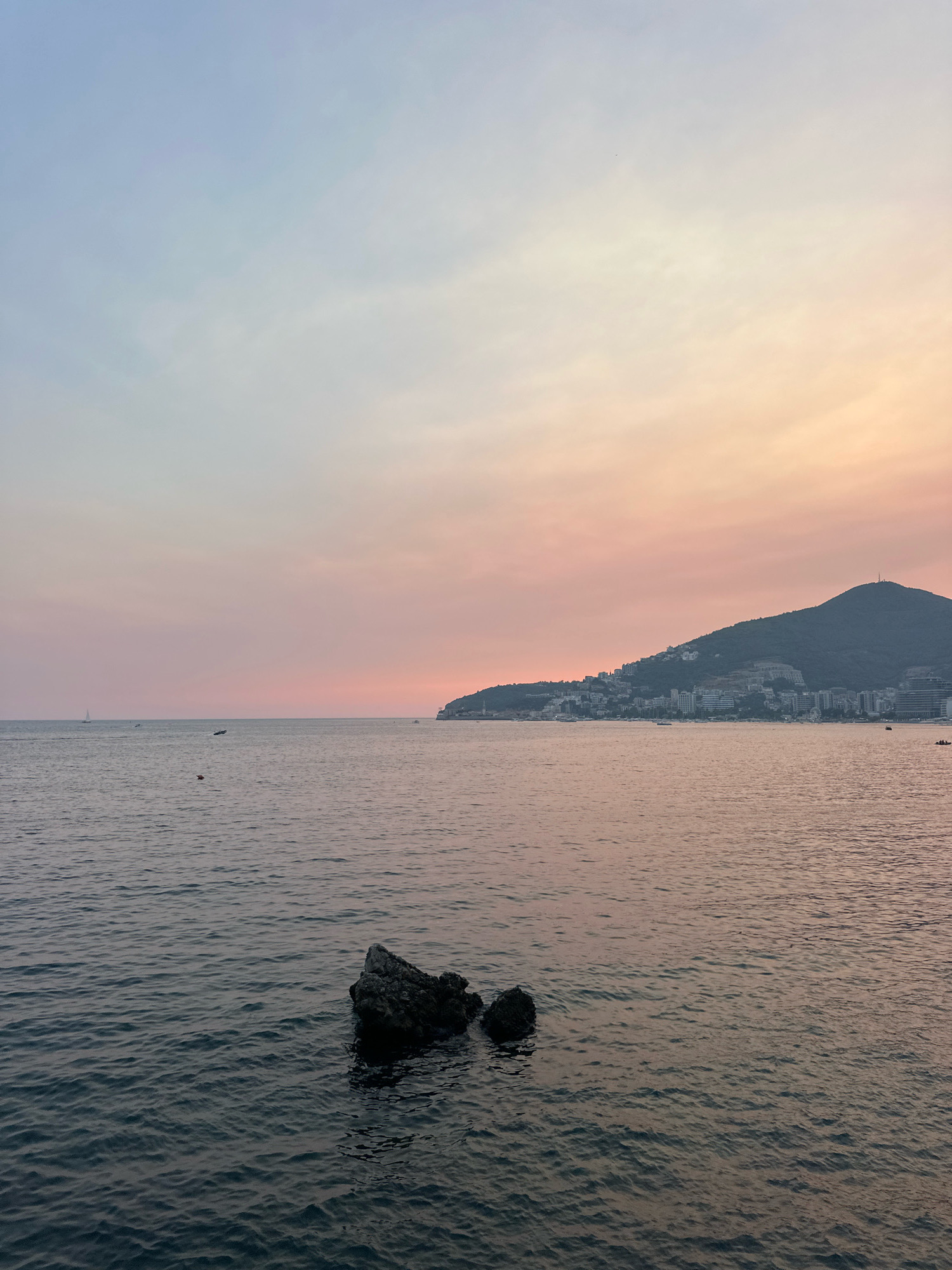 The image depicts a coastal view of Budva, Montenegro during sunset. In the foreground, dark blue ocean water shows gentle ripples and two dark rocks are partially submerged. In the midground, the calm sea stretches towards the horizon, with a few small boats visible. The background features a densely populated hillside lined with buildings and a prominent hill crowned with a structure, characteristic of Budva's urban landscape. The sky exhibits a gradient of soft pastel colors, transitioning from pale blue at the top to shades of pink and orange near the horizon, indicating the time of day. No people are directly visible.