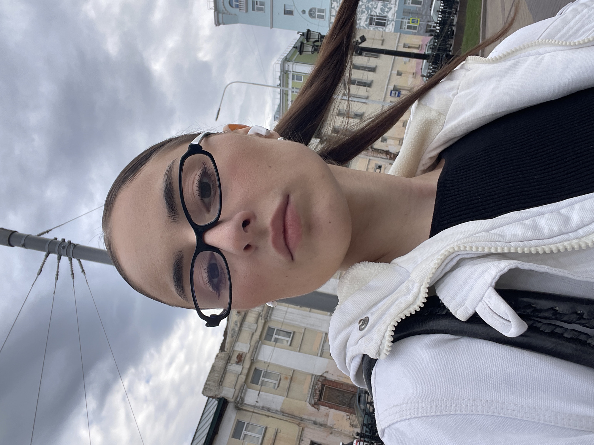A young woman with fair skin and brown hair in two high ponytails is taking a selfie. She is wearing black-framed glasses and wireless earbuds. The woman is wearing a black top and a white jacket. She is outside in an urban area with buildings in the background. Overhead wires are visible in the background. Based on the architecture, the location is likely in Moscow, Russia.