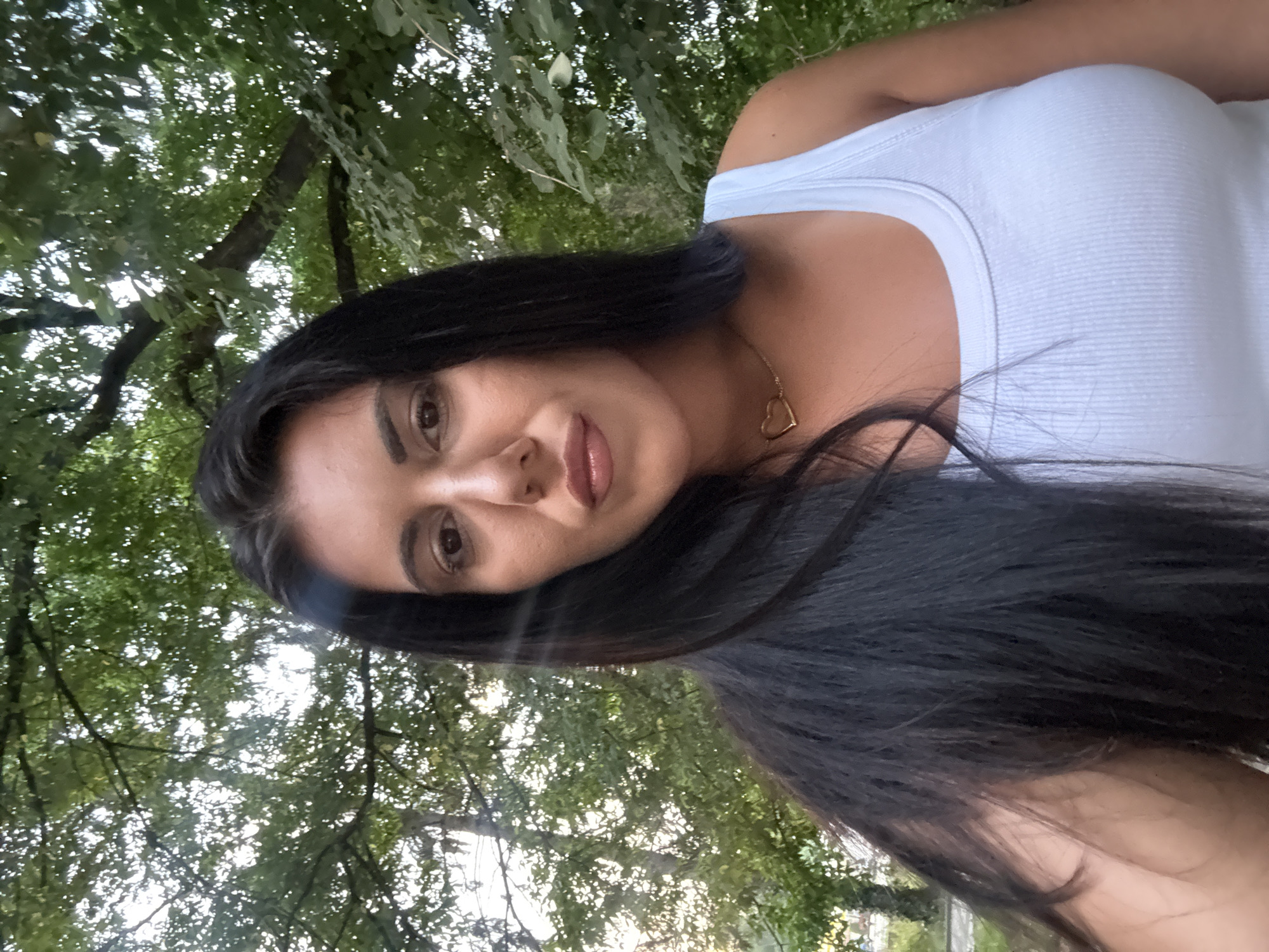 A medium-shot portrait shows a young woman with long, dark hair and a light blue tank top, wearing a gold heart-shaped necklace. She is outdoors, with lush green trees forming a natural backdrop. The lighting suggests it is late afternoon or early evening, with some dappled sunlight filtering through the leaves. The woman's gaze is directed towards the camera, and her expression is neutral. The overall impression is a casual, relaxed moment captured in a natural setting, possibly within the city of Strelcha, Bulgaria, known for its verdant landscapes.