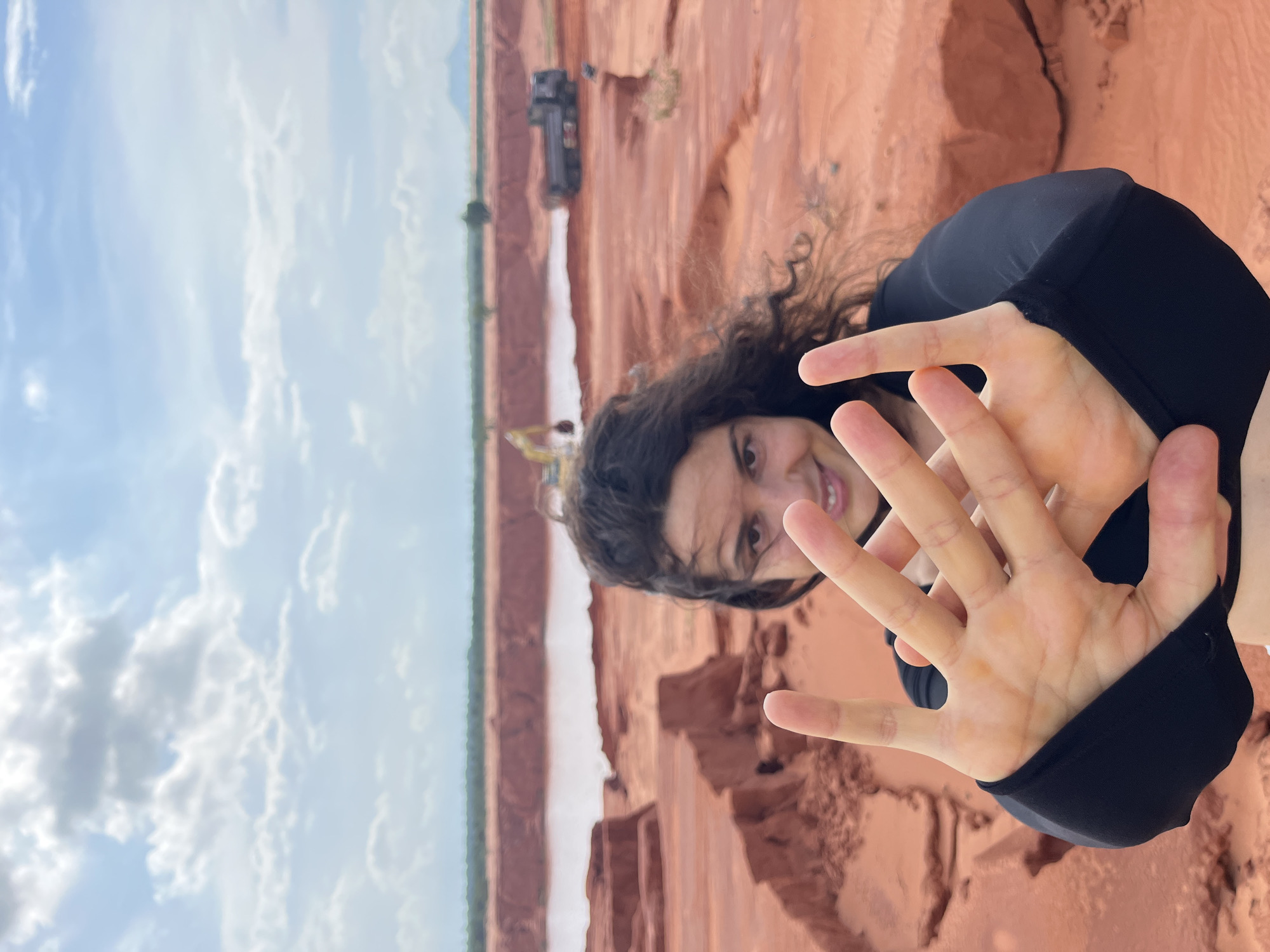 A woman with dark, curly hair and a black top is shown in the foreground, holding her hands up in front of her face and smiling. Her hands are positioned as if to block the camera or to create a frame. In the background, there is a large expanse of red earth, possibly a construction or mining site, with a body of water in the distance. An excavator and a truck are visible in the middle ground of the site. The sky is cloudy. The scene is likely a landscape with industrial or natural features.
