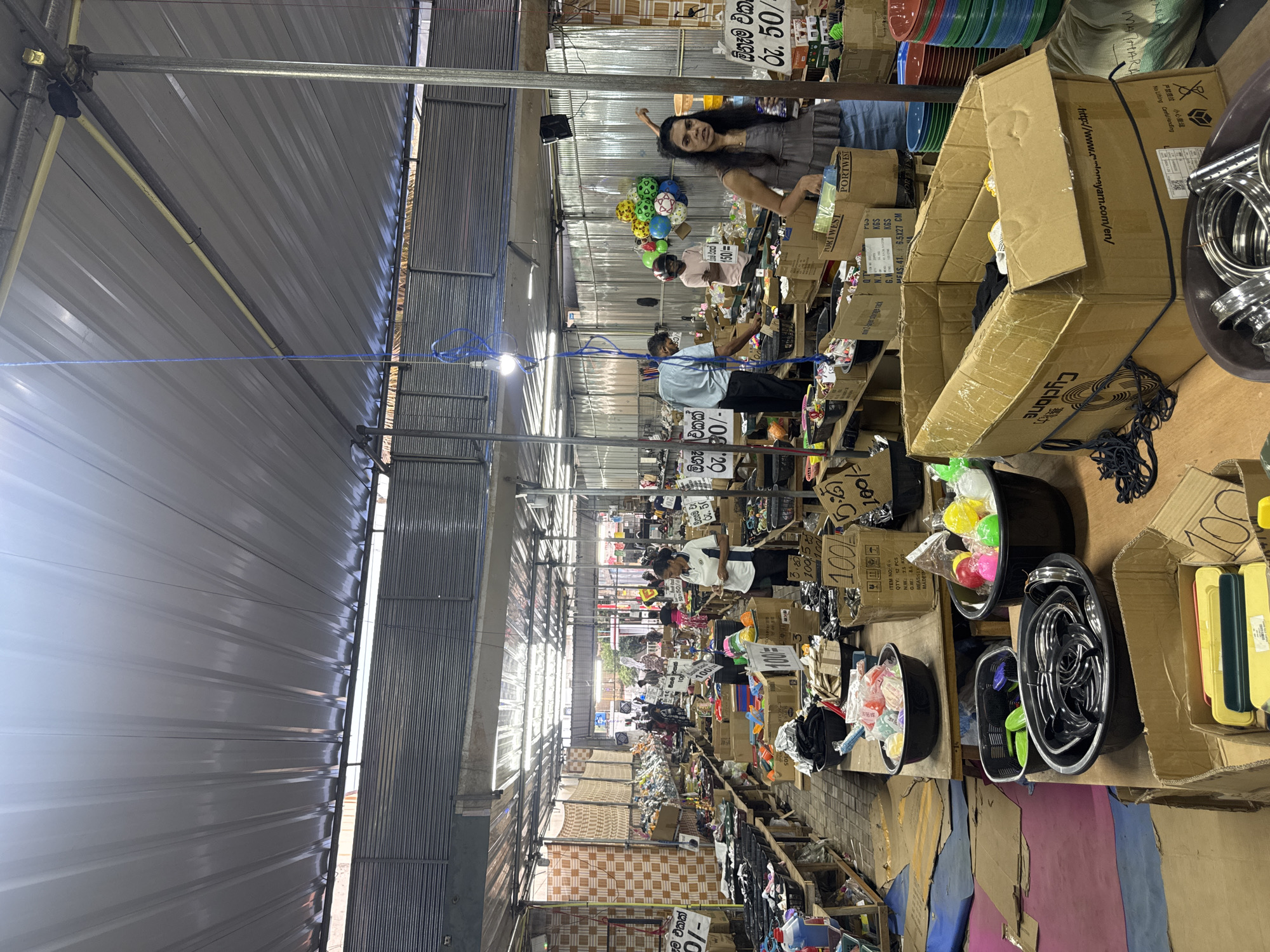 The image shows an indoor market scene in Matara, Sri Lanka. People are present, standing behind stalls filled with various items including toys, household goods, and packaged products. Many of the stalls have signs displaying prices in Sinhala. The building has a metal roof supported by metal beams. Cardboard boxes are prominent throughout the market. Lighting fixtures are suspended from the ceiling.