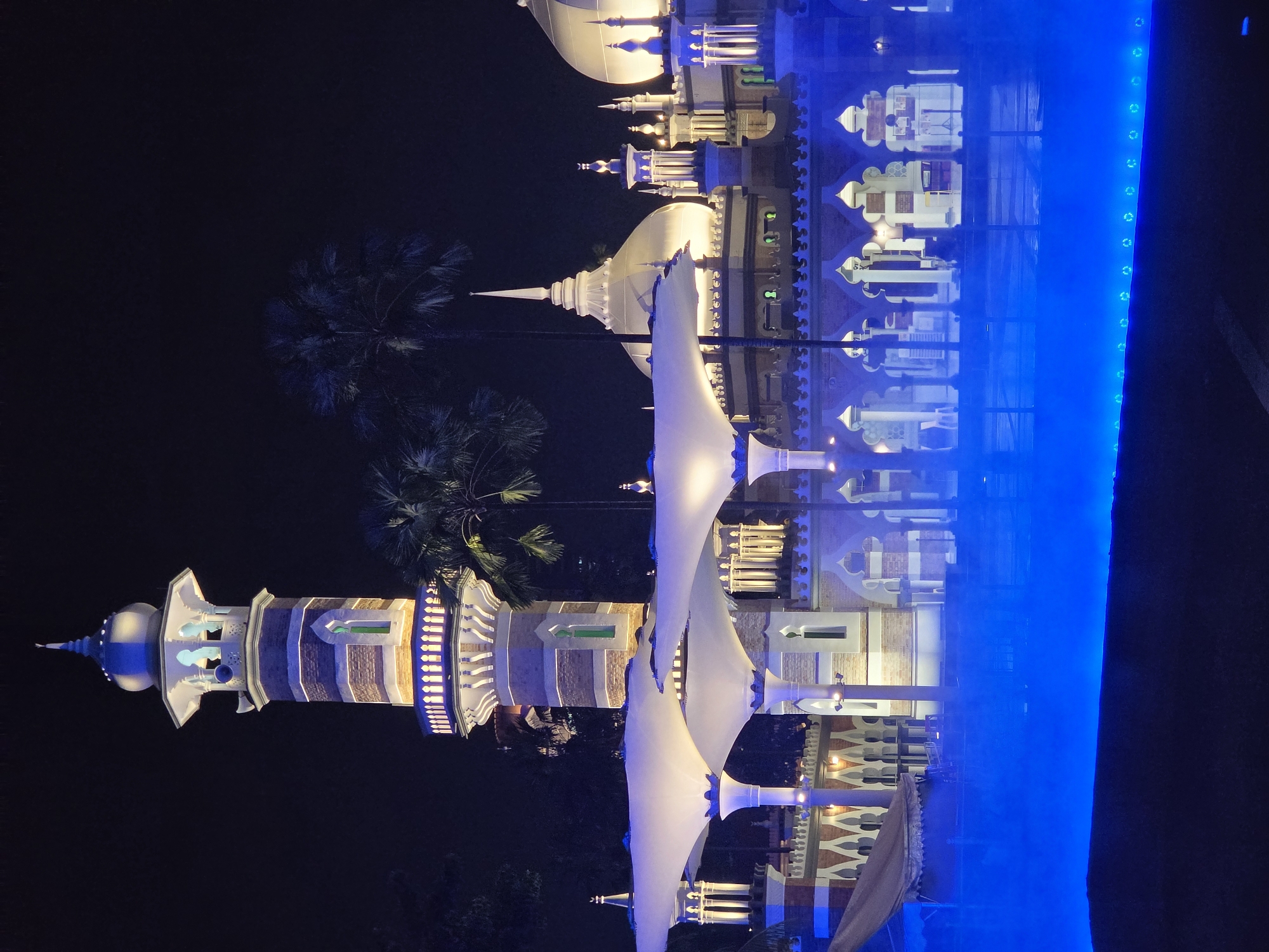 Here is a description of the image:
The image showcases a night scene in Kuala Lumpur, Malaysia, featuring the Sultan Abdul Samad Building and its reflection in a water feature. The building exhibits a blend of architectural styles and is illuminated with white and blue lights. Modern white awnings are positioned nearby. The foreground is dominated by a brightly lit blue vertical strip which is the edge of the water feature. Palm trees are visible on the left side of the image.