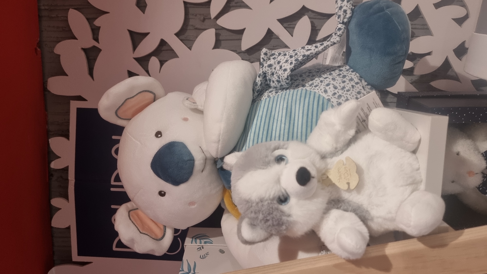 The image captures a display of baby plush toys and accessories, likely in a retail store setting in Sofia, Bulgaria.
The main subjects are three stuffed animals. Prominently featured on the left is a large white plush koala with a soft blue nose, rosy cheeks, and light pink inner ears. Its eyes are small black dots. Behind the koala's head, there's a textured fabric element, partly striped in light blue and white, and partly adorned with a blue and white floral pattern.
To the right of the koala lies a smaller grey and white plush toy resembling a husky or a wolf, featuring bright blue eyes and a black nose. A small beige tag with the partially visible text "Happy O'Day" hangs from this plush. Further to the right, a white plush bunny is visible, peeking out from a dark blue box embellished with white polka dots.
The background consists of a decorative wall with large, white, stylized leaf-like cut-outs set against a muted, possibly grey, patterned surface. A bold red vertical surface is visible on the far left. A light-colored wooden surface forms the base of the display in the foreground. Other items include a white box with a green leaf pattern on the bottom left and a dark blue sign behind the koala with some illegible white text.
The scene is brightly lit, indicative of an indoor environment, possibly a children's section or a nursery store. The arrangement suggests the items are presented for sale or as part of a product showcase.
