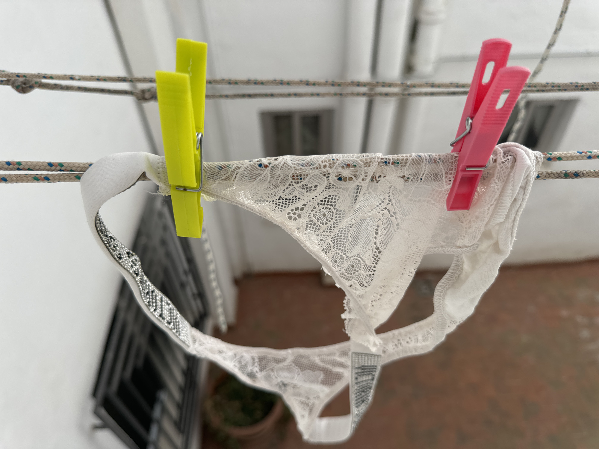 A white lace thong is hanging on a clothesline, secured by two yellow clothespins on the left and two pink clothespins on the right. The thong is made of delicate lace with a floral pattern and a white cotton gusset. The waistband appears to have a grey and black patterned texture. The background shows an outdoor setting with white walls and hints of windows. Below the clothesline, a terracotta-colored surface can be seen, possibly a patio or balcony. A potted plant is partially visible in the lower left corner. The scene suggests a sunny day, given the bright lighting. The location is identified as Murcia, Spain. There is no visible text in the image.