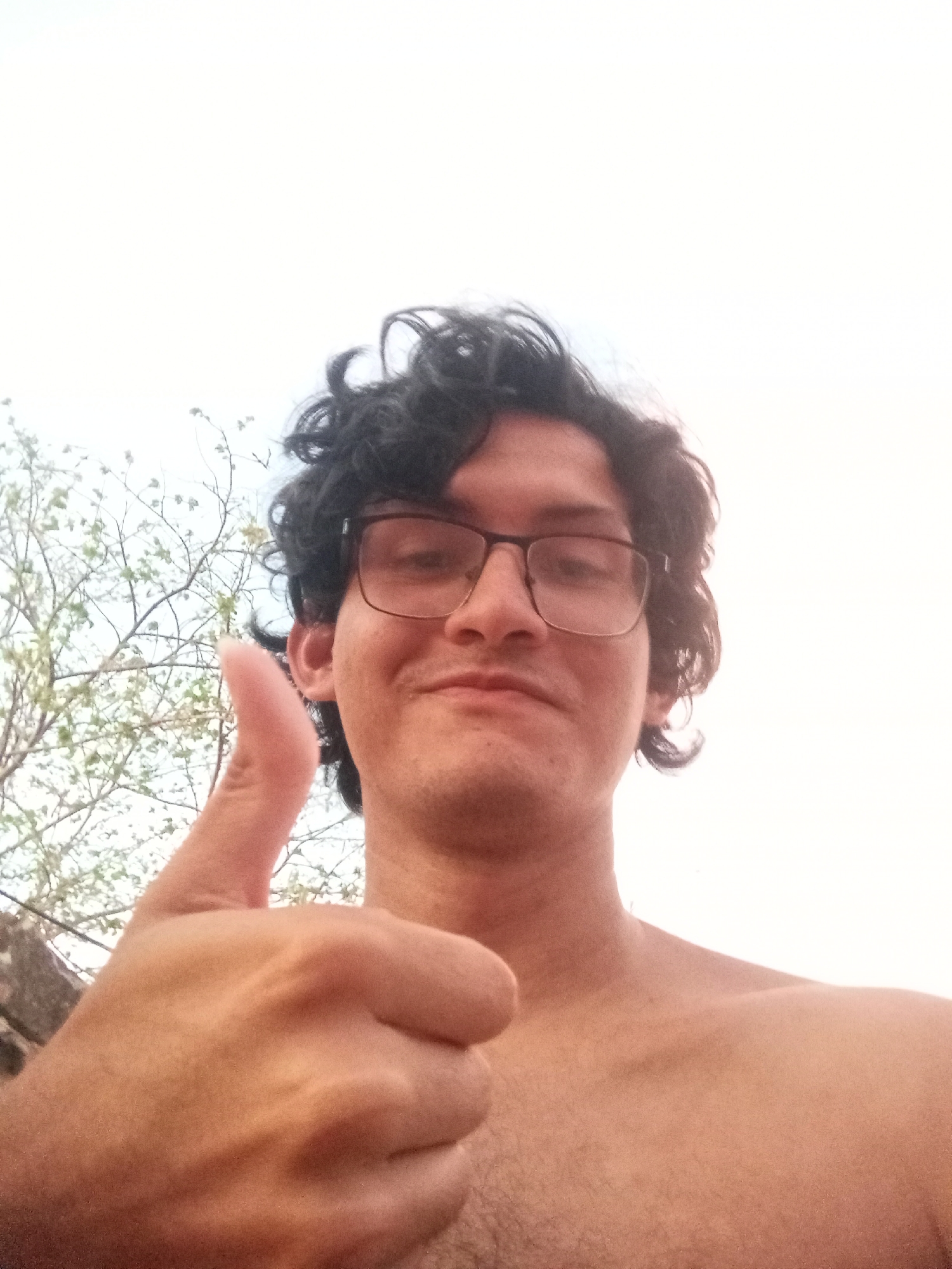 A selfie-style, upward-angle photograph depicts a man with dark, curly hair and glasses giving a thumbs-up gesture. The man is shirtless, revealing his upper chest and shoulders. A tree with budding leaves is visible behind him and to the left. The backdrop is a bright, overcast sky, suggesting an outdoor setting. The location is likely Juan Griego, Venezuela based on the description.