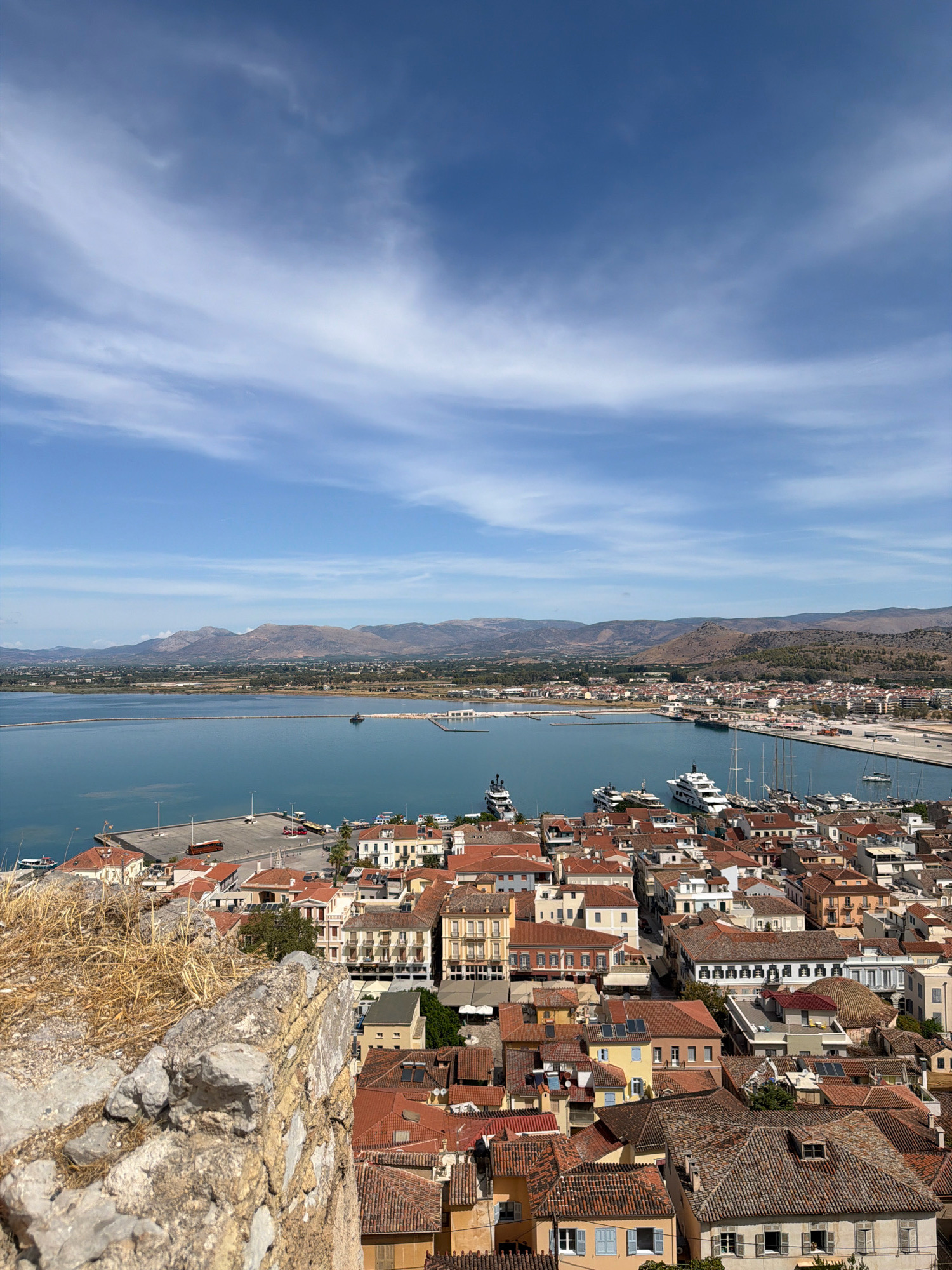 A vertical, medium-shot, elevated perspective captures a sunlit coastal town under a bright blue sky with wispy white clouds. The foreground features a weathered, stone wall, partially covered in dry, yellow grass. Beyond the wall, a dense collection of buildings with terracotta tiled roofs stretches across the landscape. The architecture appears traditional, with cream, ochre, and white facades.
The middle ground is dominated by a calm, blue body of water, likely a harbor. Several boats, including large white yachts, are docked or moored in the harbor. A paved quay runs along the edge of the water, with a few vehicles visible.
In the background, a distant, mountainous horizon is visible. The overall scene depicts a scenic view of the Municipal Unit of Nafplio, Greece, likely from a vantage point like a castle or elevated historical site, offering a panoramic vista of the town, harbor, and natural landscape. The image is devoid of any people.