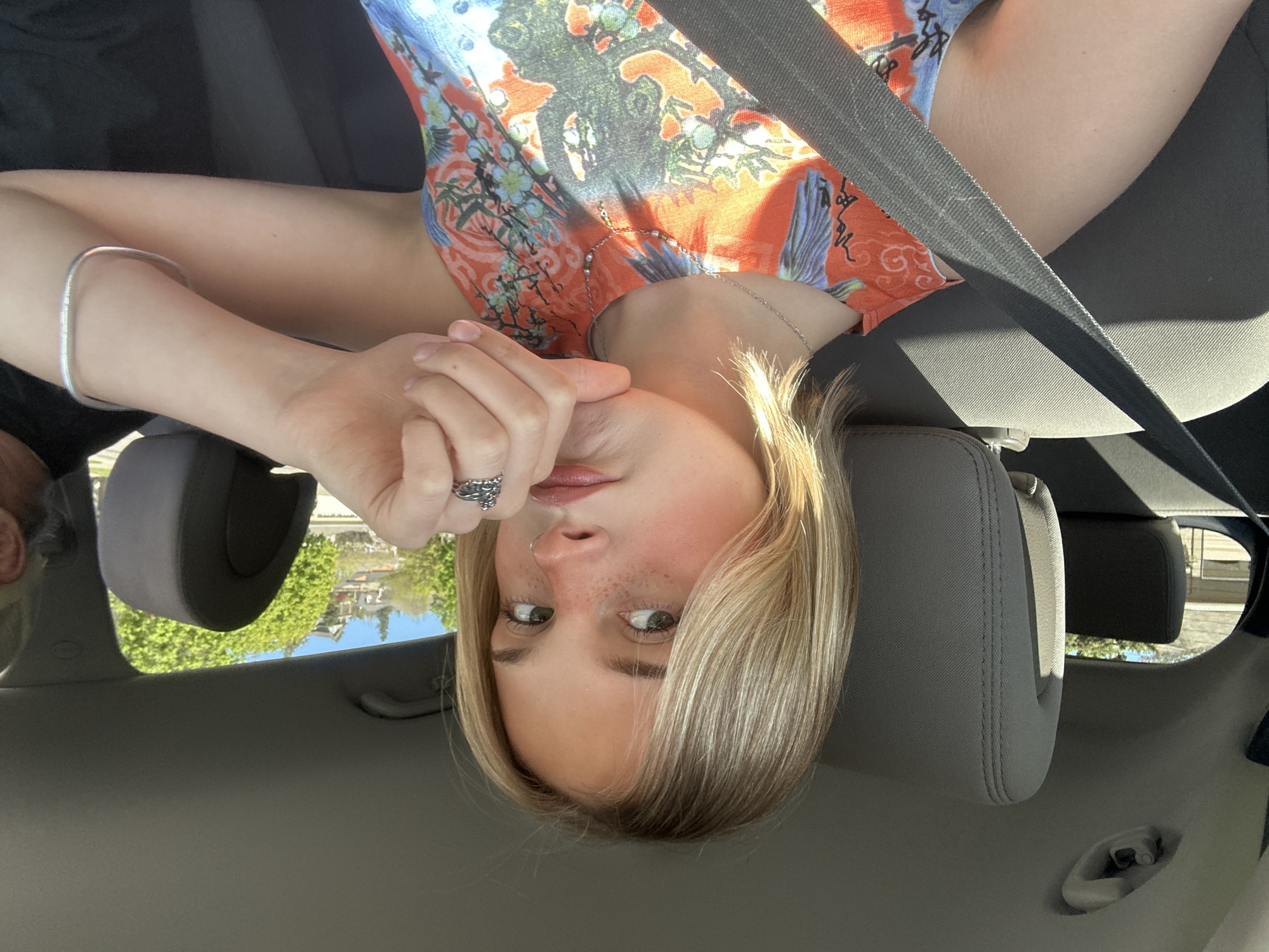 A selfie photograph shows a woman with blonde hair and freckles in the front passenger seat of a vehicle. She is wearing a colorful patterned shirt, a silver bracelet, and a ring. A black seatbelt crosses her body. The background outside the car window shows trees and houses, suggesting a residential area, but no specific landmarks in Kelowna are identifiable.