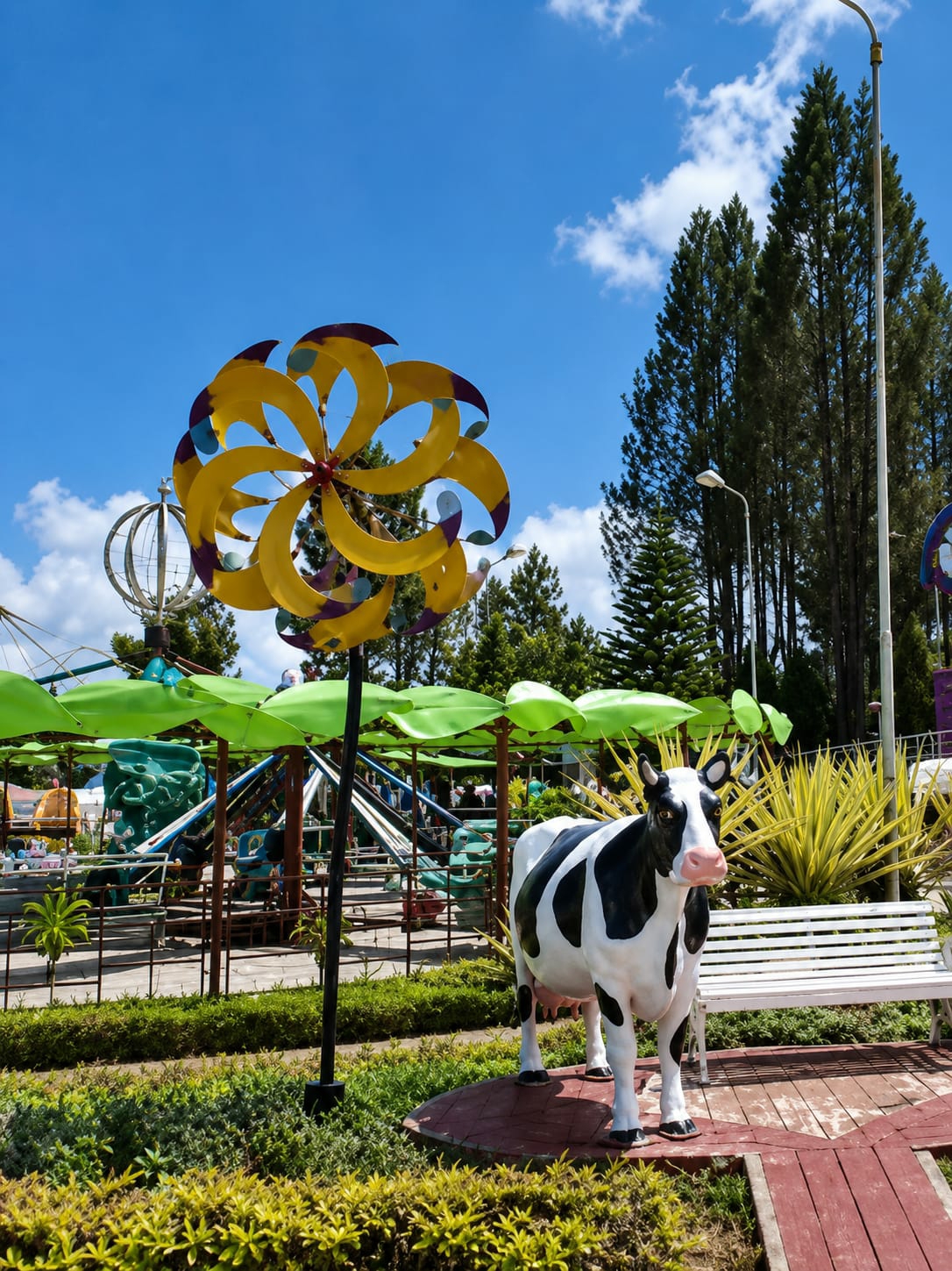 The image depicts an outdoor scene in what appears to be a park or amusement area, likely in Berastagi, Indonesia, given the context. In the foreground, a life-sized, realistic statue of a black and white cow stands on a red circular platform. Behind the cow, a white slatted park bench is visible. To the left of the cow, a large, colorful pinwheel made of yellow and purple curved blades spins on a black pole. In the middle ground, there is an amusement ride with green leaf-like canopies and various colored seats and structures. Beyond the ride, tall, dark green coniferous trees line the background, reaching towards a bright blue sky with scattered white clouds. The environment is lush with green bushes and manicured hedges in the foreground and middle ground. The lighting suggests a sunny day.
