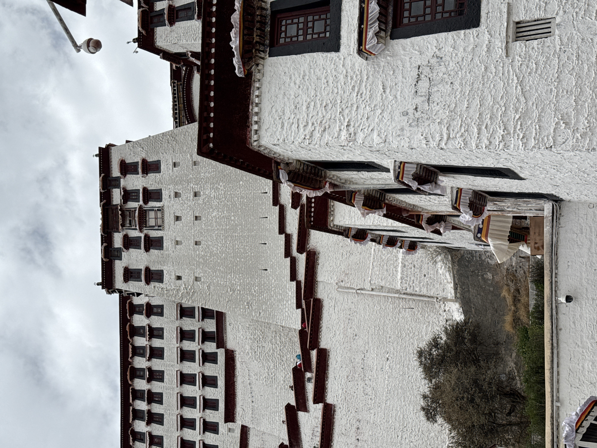 The image depicts a section of the Potala Palace in the Chengguan District of Lhasa, Tibet Autonomous Region, China. The architecture features thick white walls with numerous windows outlined in dark trim. Decorative maroon-colored accents and draping fabric are visible along the roofline. The sky is overcast. A tree is visible in the foreground, and a security camera is affixed to a wall. The image suggests an exterior shot focused on the architectural details of the palace.