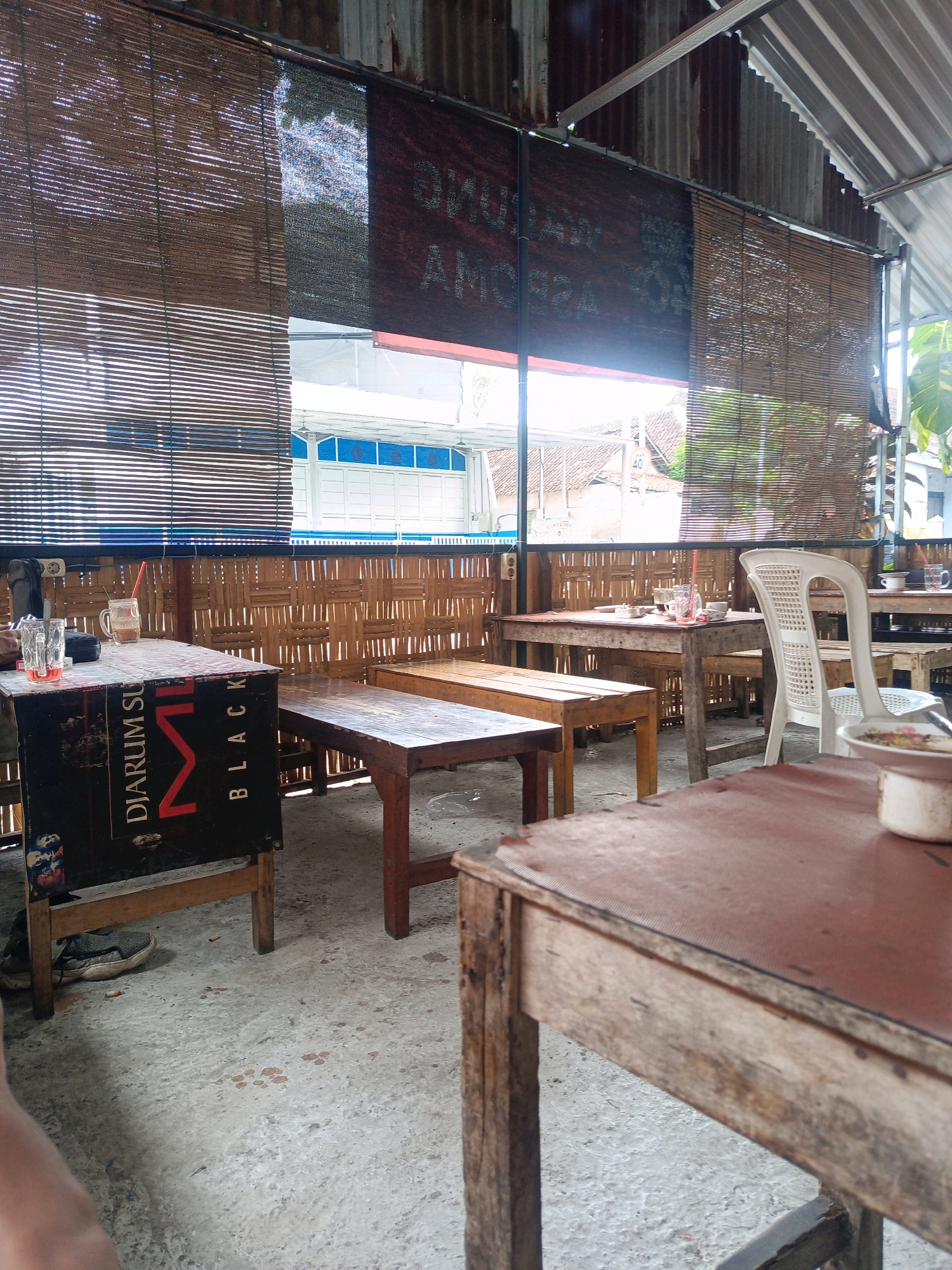 The image depicts an outdoor seating area, likely a warung or small restaurant, in Magetan, Indonesia. The scene is characterized by a rustic, open-air ambiance with a corrugated metal roof and bamboo-woven walls. Wooden tables and benches are arranged on a concrete floor. In the foreground, a wooden table with a reddish-brown top is prominently featured. To the left, a black box with red lettering, possibly a cigarette advertisement for "DJARUM SUPER BLACK," serves as a makeshift table or stand. Two glasses, one with a straw and a reddish liquid, and another with a clear liquid, sit on this box. A white plastic chair is positioned at a table in the mid-ground. Behind the tables, bamboo blinds hang as window coverings, allowing sunlight to filter through. A glimpse of buildings and vegetation is visible through the blinds. The overall impression is that of a casual, informal dining or resting spot.
