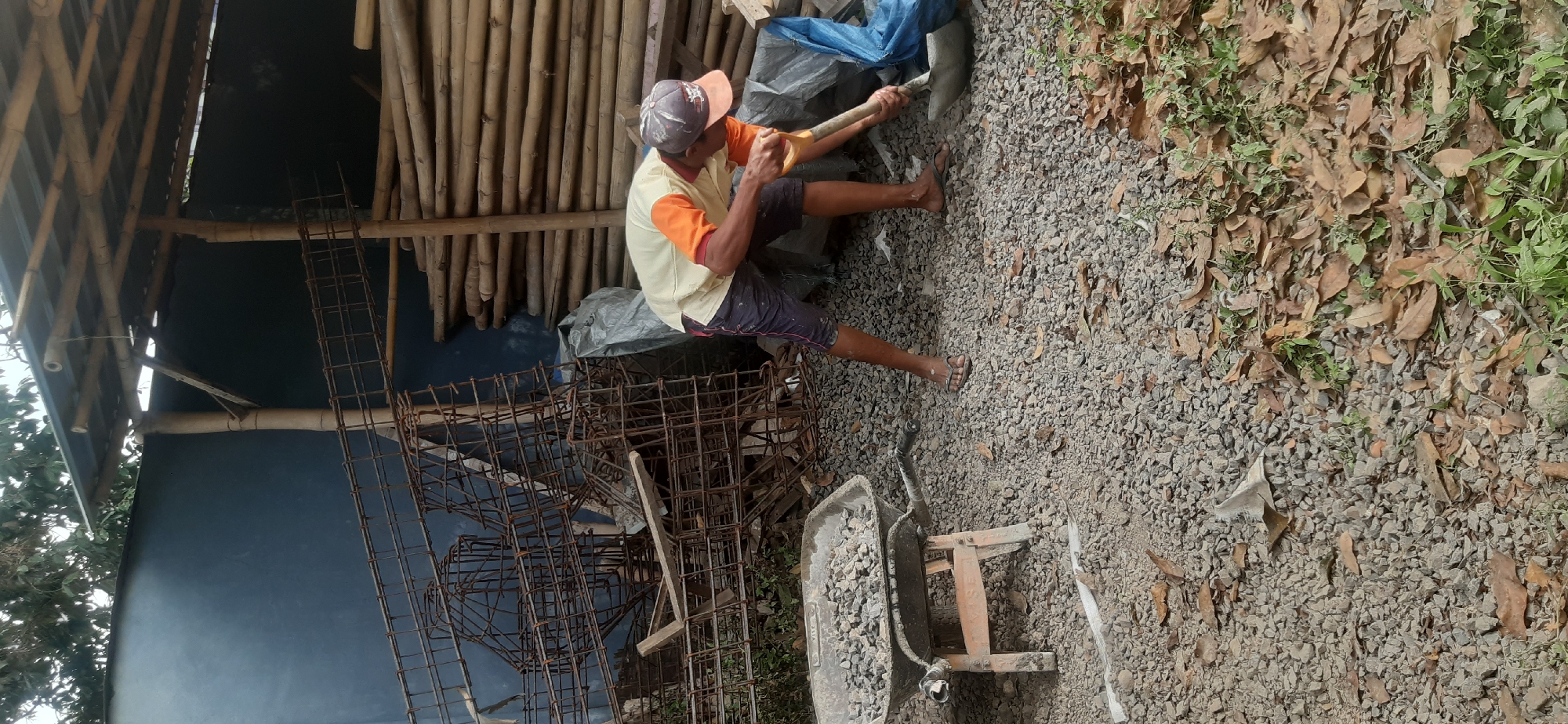 A lone construction worker, wearing a light yellow t-shirt with orange accents and dark shorts, is actively working on a construction site. He is using a yellow shovel to mix what appears to be cement in a blue tarp. The ground is covered with small grey stones, and fallen brown leaves are scattered around. In the background, there is a pile of stacked bamboo poles, a dark blue wall, and some metal rebar structures. A wheelbarrow filled with grey gravel is positioned to the left of the worker. The scene suggests an outdoor construction or renovation project in a rural or semi-rural area, possibly in Sepakek, Indonesia, given the materials and building style.