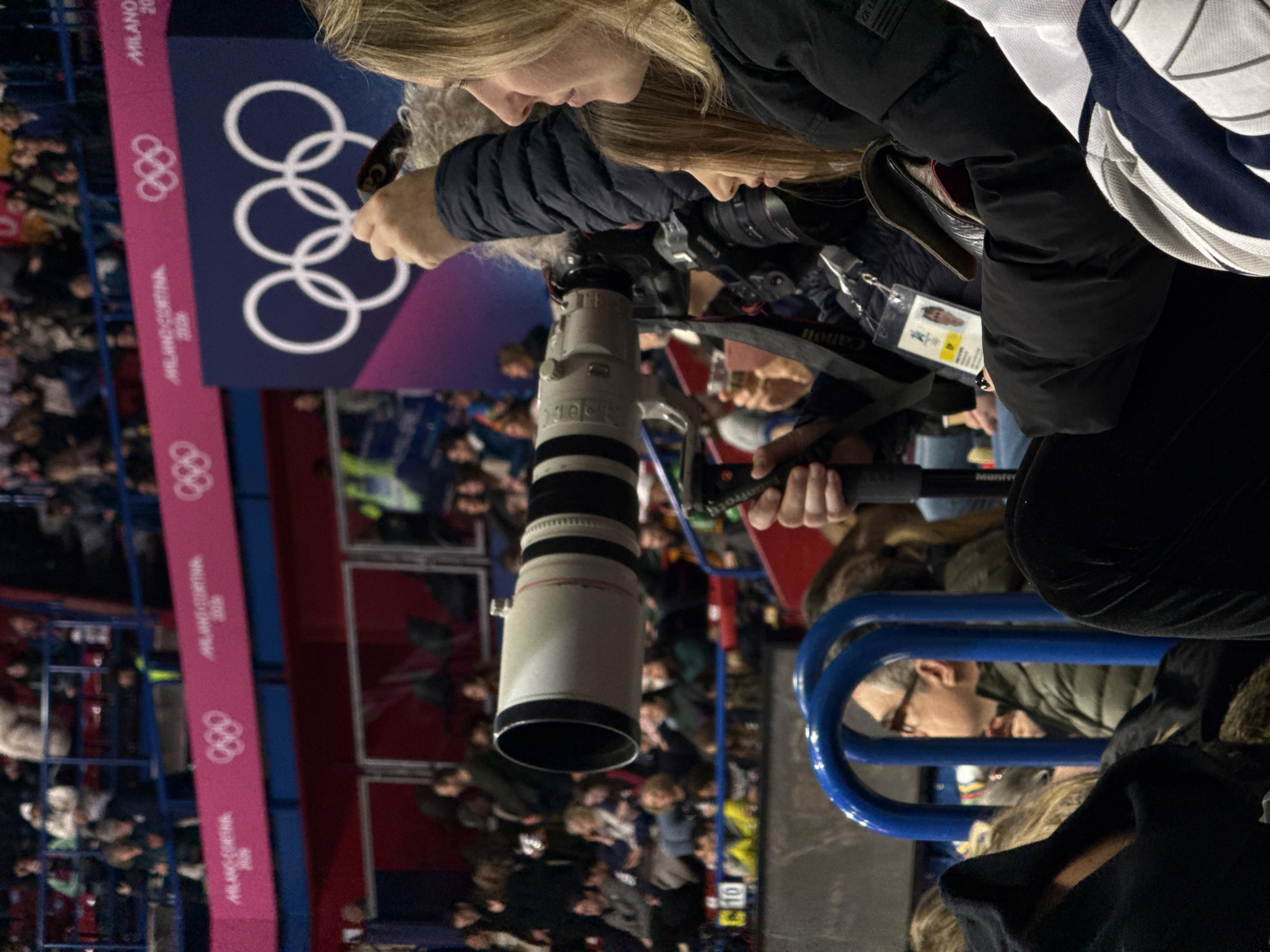 The image captures a close-up view of a photographer at what appears to be an Olympic event. In the foreground, a woman with blonde hair is seen from the side, looking downwards with a focused expression. Her hands are positioned on a large, professional-grade telephoto lens, which is predominantly white with black rings, indicating a high-end camera setup. A camera strap with the brand "Canon" is visible near her.
To her right, a black handle or support for the camera, possibly part of a monopod or tripod, bears the brand name "MANFROTTO". An ID badge on a lanyard hangs around her neck, though the details on it are mostly obscured, except for a small portrait image. She is wearing a dark, possibly padded, jacket.
In the mid-ground and background, the setting is an indoor arena or stadium, densely populated with spectators who are mostly blurred, indicating depth of field focusing on the foreground subjects. Prominently displayed in the background are banners or signs featuring the Olympic rings logo. The text "MILANO CORTINA 2026" is clearly visible multiple times on these banners, indicating that the event is related to the 2026 Winter Olympics in Milan and Cortina d'Ampezzo. The banners are colored in shades of dark blue/purple and pink.
A blue metal railing is visible in the lower right and center of the frame, separating the viewing area where the photographer is located from what appears to be another section of the arena or audience. Below this railing, part of another person with gray hair and glasses can be seen. The overall lighting suggests an indoor event, likely under artificial illumination. The image is taken from a slightly elevated position, looking down towards the event space and other spectators.