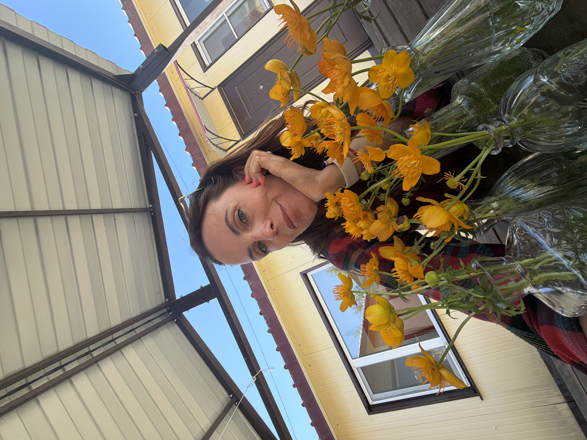 A woman is posing for a selfie outdoors, possibly on a porch or patio, with a bright yellow building and a blue sky visible in the background. She is resting her head on her hand, looking at the camera. In the foreground, there are several glass vases filled with orange flowers. The woman is wearing a plaid scarf. The setting appears to be a residential area, and given the mention of Birobidzhan, Russia, it's likely a backyard or a similar outdoor space in that city.