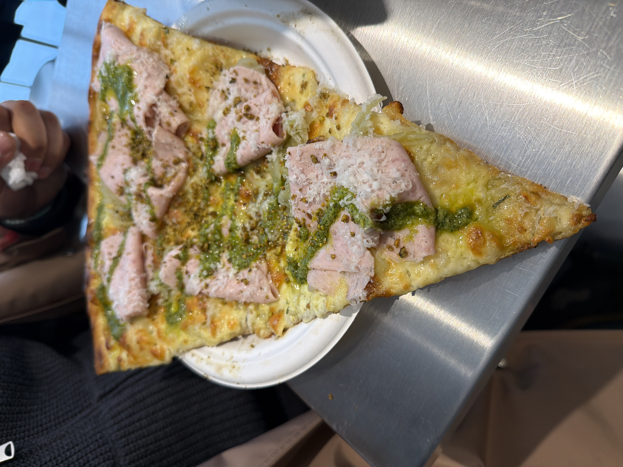 A slice of pizza sits on a white paper plate, placed on a stainless steel surface. The pizza has a pale crust topped with cheese, slices of pale pink meat, and a green pesto-like sauce. Grated cheese is sprinkled on top of the meat. The pizza slice appears to be partially consumed, with a bite taken from the pointed end. A hand holding a crumpled white napkin is visible in the upper left corner. The location appears to be in Unknown, Unknown.