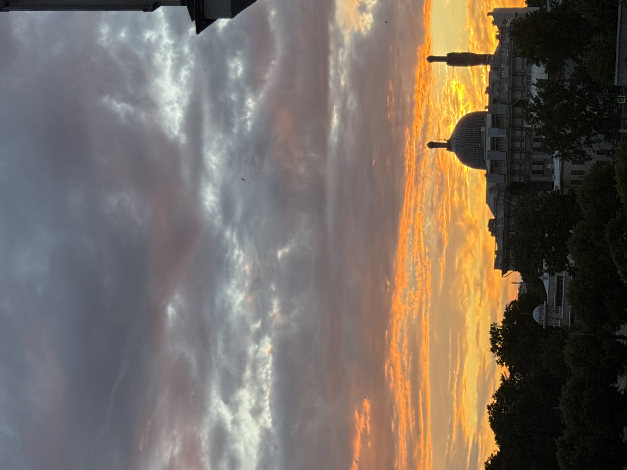 The image captures a dramatic sunset over Saint Petersburg, Russia. The sky is filled with a striking gradient of colors, transitioning from deep purples and grays on the left to vibrant oranges and yellows on the right, illuminated by the setting sun. Wispy clouds are painted with the warm hues of twilight, creating a textured and dynamic celestial display.
Silhouetted against the fiery sky are the architectural elements of the city. A domed building, characteristic of Saint Petersburg's historical architecture, is clearly visible. A tall, slender monument or spire also stands prominently, adding to the urban skyline. The foreground is partially occupied by the dark mass of trees, framing the view of the buildings and the sky. The overall scene evokes a sense of awe and tranquility, common emotions associated with witnessing a beautiful sunset. The time of day is clearly evening, during the golden hour. No visible text is present in the image.