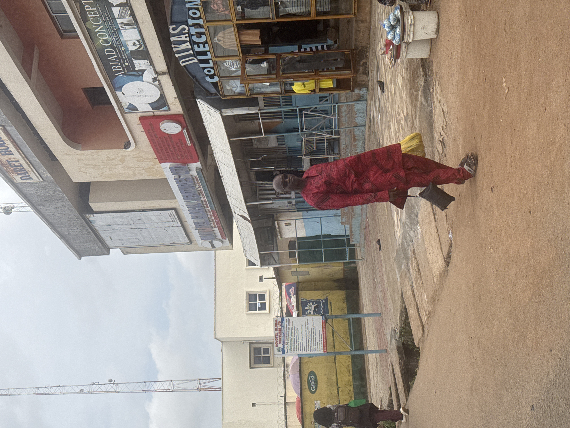 The image shows a street scene in Jos, Nigeria. A man in a red and black patterned traditional outfit is walking on a sandy, uneven ground. He is carrying a yellow bag over his shoulder and a black bag in his hand. In the background, there are shops with displays of clothing. One shop has a prominent sign that reads "DIKAS COLLECTION". Another sign, partially visible, says "ABIAD CONCEPT". Further down the street, there are more buildings and signs. A large crane is visible in the distance, suggesting ongoing construction or infrastructure work. The sky is overcast, indicating a cloudy day. The overall impression is of a busy marketplace or commercial area.