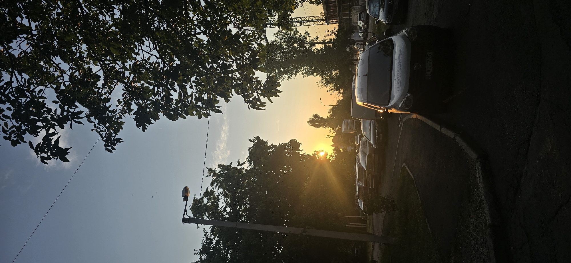The image depicts a street scene in Chișinău, Moldova, during sunset. The sun is low on the horizon, casting a warm glow and long shadows. A streetlamp is visible on the left, and trees with dense foliage frame the scene. A line of parked cars is situated on the right side of the road, with a dark-colored van in the foreground. In the background, a construction crane and other vehicles are discernible. The overall impression is one of an urban environment at dusk.