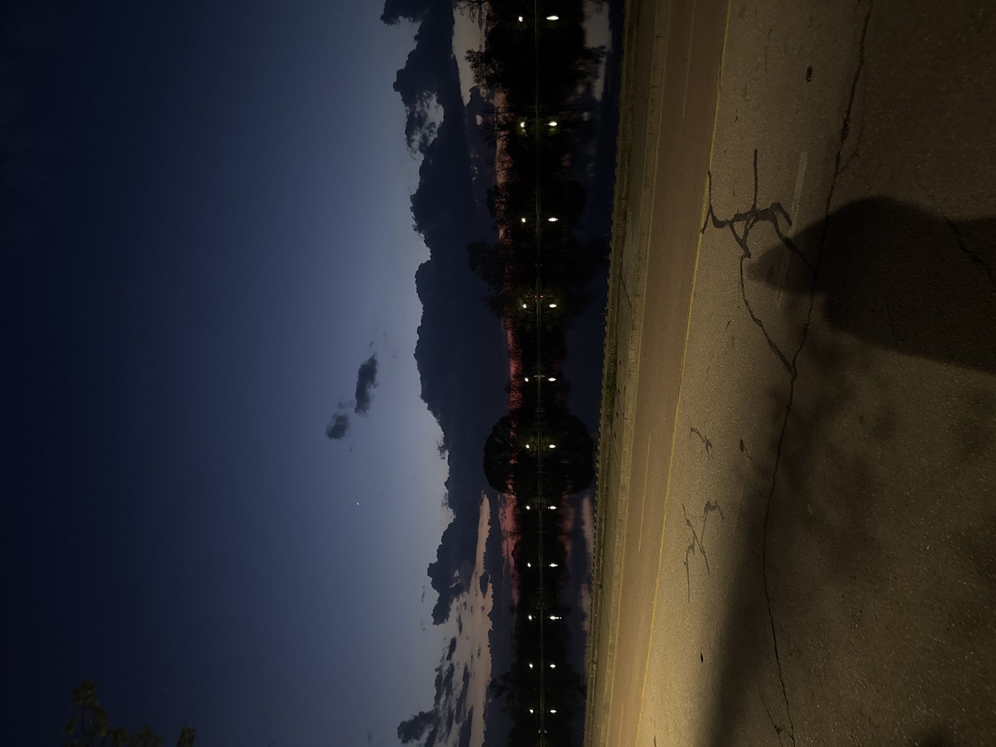 The image shows a night scene in Plovdiv, Bulgaria, likely near a body of water. The left side of the frame features a dark blue sky with scattered clouds, some reflecting pink hues from the setting sun. A crescent moon is visible. The central portion of the image showcases the reflection of trees and streetlights in what appears to be a lake or river. A line of evenly spaced lights is reflected in the water, creating a symmetrical pattern. To the right, a paved road or walkway is illuminated by the streetlights, displaying cracks and a visible shadow cast by an unseen figure or object. A yellow line separates the road from the water's edge.