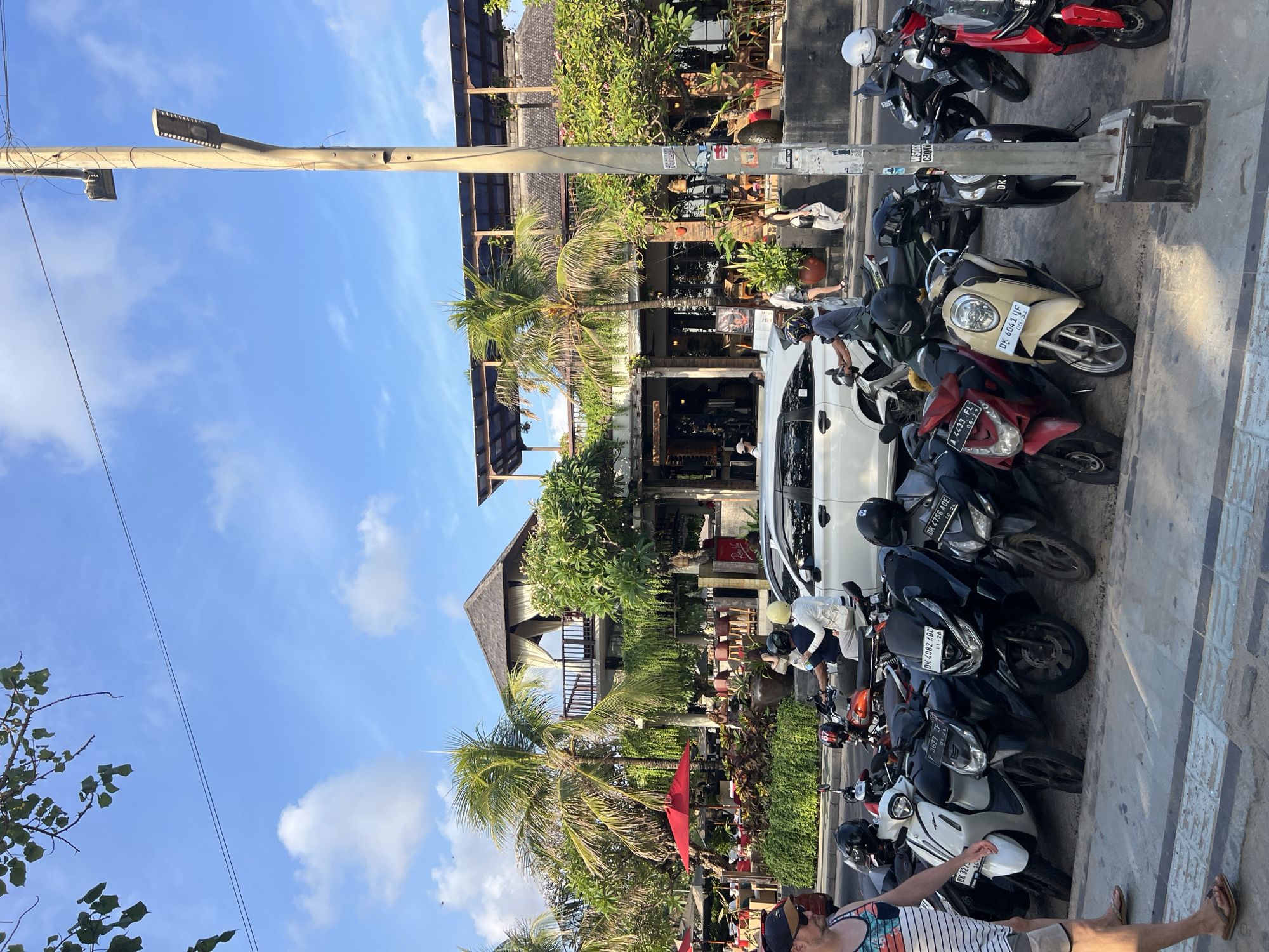This image depicts a street scene in Kuta, Indonesia, characterized by a row of parked motorcycles and scooters along a sidewalk. In the background, a multi-story building with a thatched roof and open-air balconies suggests a restaurant or commercial establishment. Tropical palm trees and lush greenery are visible, contributing to the tropical ambiance. Several people are present, including individuals walking on the sidewalk, riding motorcycles, and sitting in the background establishment. The weather appears to be clear with a blue sky and scattered clouds. The overall scene portrays a typical day in a bustling tourist area.