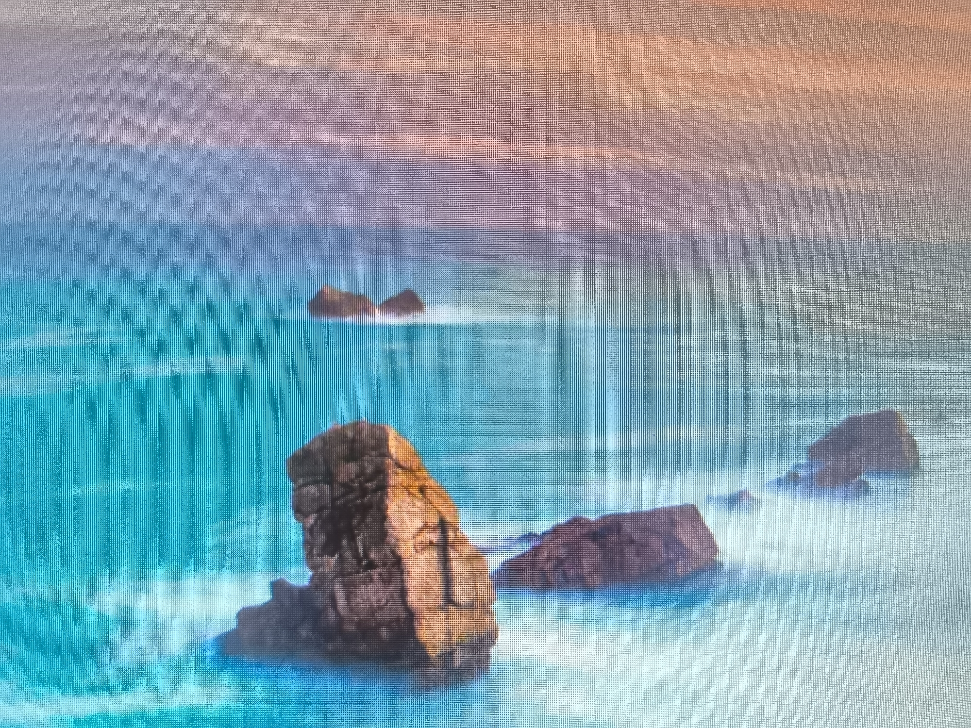 This media file depicts a serene seascape with prominent rock formations. The scene is characterized by the vibrant turquoise water crashing against dark, rugged rocks. In the foreground, a large, textured rock is illuminated by warm light, suggesting it is either sunrise or sunset. Further out in the water, several other rocky outcrops are visible, some partially submerged by the foamy waves. The water itself appears dynamic, with white froth and streaks indicating movement and the force of the ocean. The sky above is a gradient of soft colors, transitioning from a warm orange at the top to shades of pink and blue, further enhancing the impression of either dawn or dusk. The overall mood is tranquil and majestic, showcasing the raw beauty of nature. There is no visible text in the image.