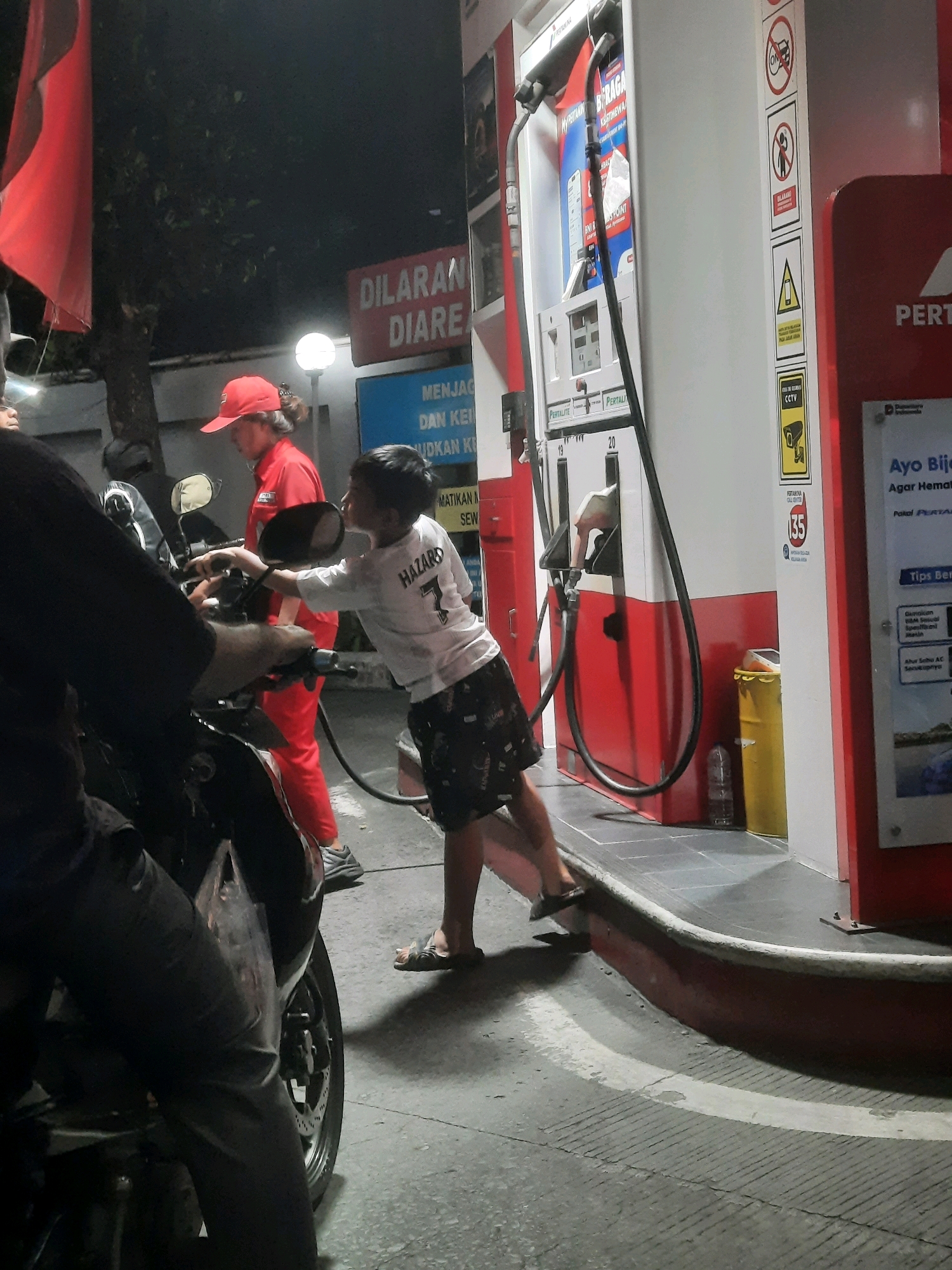 A young boy in a white t-shirt with "HAZARD 7" printed on the back and patterned shorts is standing at a gas station, reaching towards a motorcycle. He is leaning on the motorcycle's handlebars while looking at the gas pump. A person wearing a red uniform and a red cap, likely a gas station attendant, is standing behind the boy, looking down. The gas station appears to be Pertamina, a brand common in Indonesia. The scene takes place at night, with artificial lighting illuminating the area. The ground is paved, and there are warning signs and promotional posters visible on the gas pump and nearby structures. A red flag is visible on the left side of the image. The overall impression is a casual refueling stop at a busy, well-lit gas station.