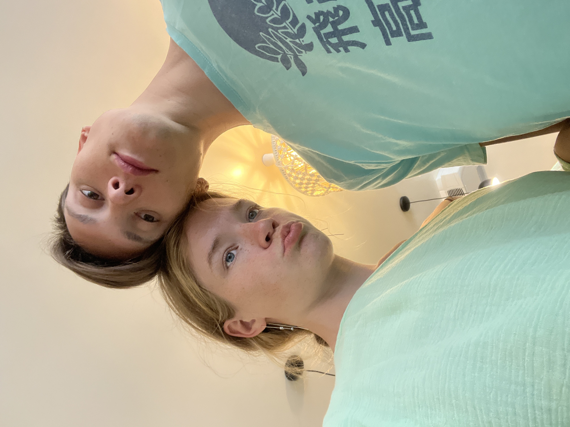 In Lapčići, Montenegro, two young individuals, a male and a female, are captured in a low-angle, close-up selfie. The female, positioned on the left, has blonde hair and blue eyes, with her head tilted and lips puckered. She is wearing a light mint green top. The male, on the right, has brown hair and is leaning his head against the female's. He is wearing a teal t-shirt with dark blue Japanese characters on it. The background is a light-colored wall with a decorative lamp visible on the right side of the frame. The lighting suggests an indoor setting, possibly a room with ambient light from a lamp.