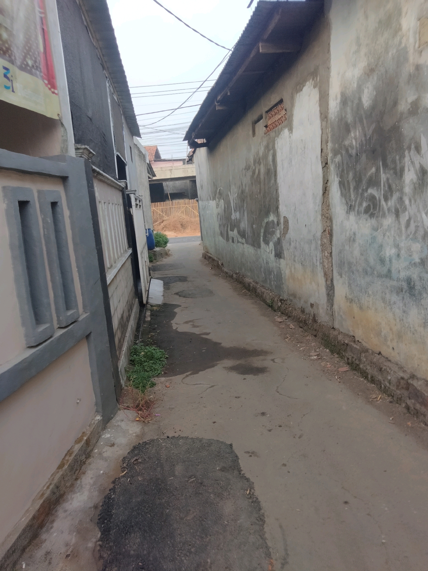 The image depicts a narrow, paved alleyway in Suka Sari, Indonesia. The alley is bordered by two buildings. On the left, a light-colored building with a grey fence is visible, featuring a large banner with Indonesian text and blue graphics. The fence has vertical decorative elements. On the right, a weathered, grey concrete wall with graffiti covers a significant portion of the image. The wall shows signs of decay and has a section of exposed brickwork. Overhead, electrical wires are strung across the sky. The ground of the alley is asphalt, with some patches of dark, wet-looking areas and debris. To the far right of the alley, a rustic wooden fence encloses an area with dried vegetation. The scene suggests an urban or semi-urban residential area, possibly in a developing neighborhood, given the condition of the buildings and the unpaved areas visible in the distance.