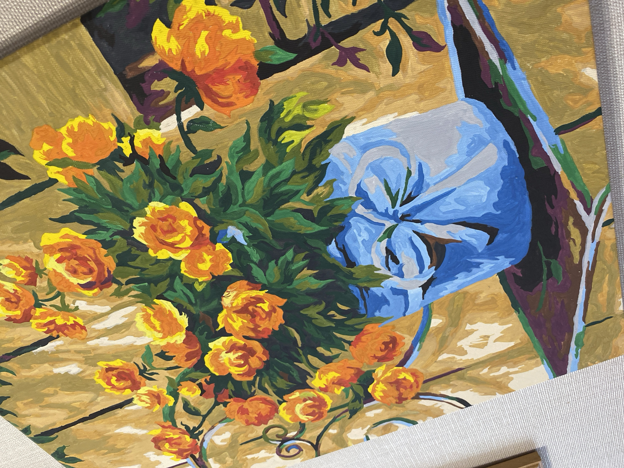 The image depicts a close-up view of a painting. The painting features a still life arrangement with yellow and orange flowers, predominantly roses, in a vase. The vase is a blue color, appearing to be tied at the top with a gathered cloth or similar material. The background is a golden hue, suggesting a table surface or wall. There are dark brown and green stems and foliage accompanying the flowers. The painting style is somewhat stylized and possibly amateur in execution. The painting is framed in a light-colored, textured frame. There is no information available in the image or the given prompt to identify the location as Kauchuk Stantsiya, Russia. There are no people visible or suggested by the context of the image.