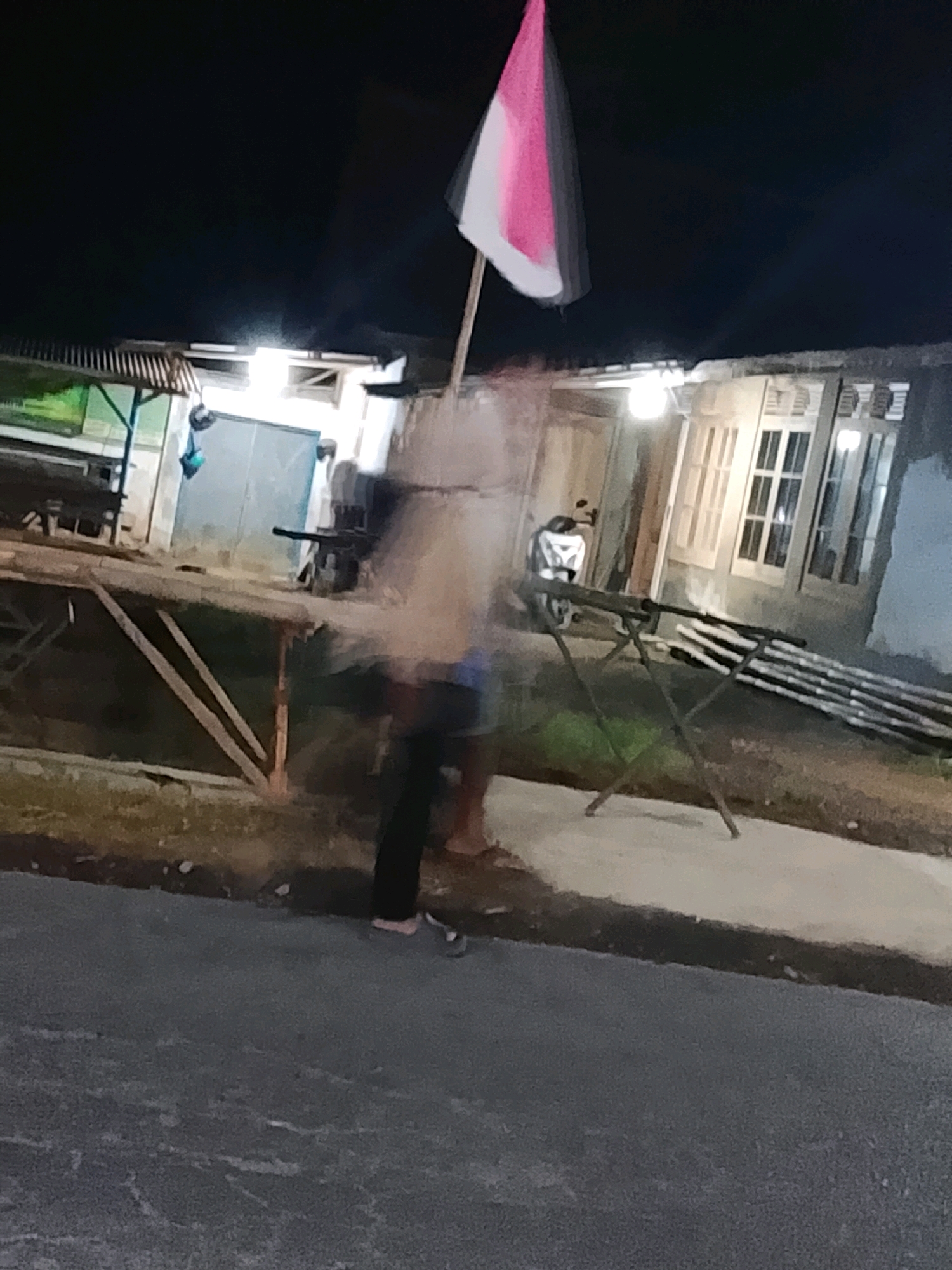 The image depicts an outdoor scene at night, likely in Sukorejo, Indonesia. In the foreground, a blurred figure, possibly a person, is visible on a paved surface. To the left, a wooden structure with diagonal supports extends into the scene. Behind this, a building with several windows is illuminated. A motorcycle is parked to the left of the building entrance. A flagpole with a red and white flag, appearing blurred and distorted, is positioned centrally, extending upwards. The overall lighting suggests artificial illumination from the building.