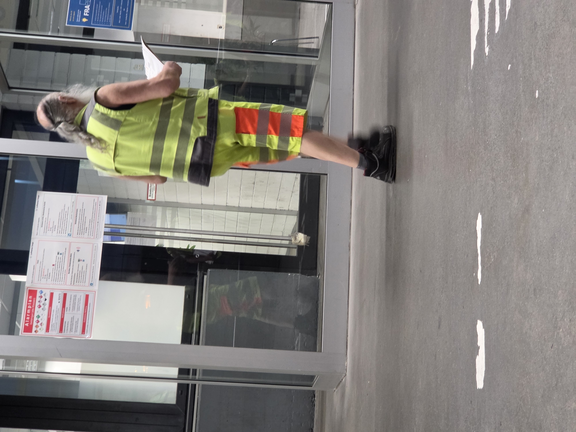 The image depicts a man wearing a high-visibility yellow vest with reflective stripes and yellow shorts with orange reflective stripes, along with black work boots. He is holding a white sheet of paper in his right hand. The man has long grey hair tied back in a braid and a beard. He is walking on a grey asphalt surface. In the background, there is a glass building facade with posters displayed on the interior wall, one of which is labeled "Alarmplan". There are also reflections in the glass, including a glimpse of what appears to be a dark colored dog. The scene appears to be in an urban outdoor environment. The presence of the "FRA" logo on a poster in the background suggests a location at Frankfurt Airport.