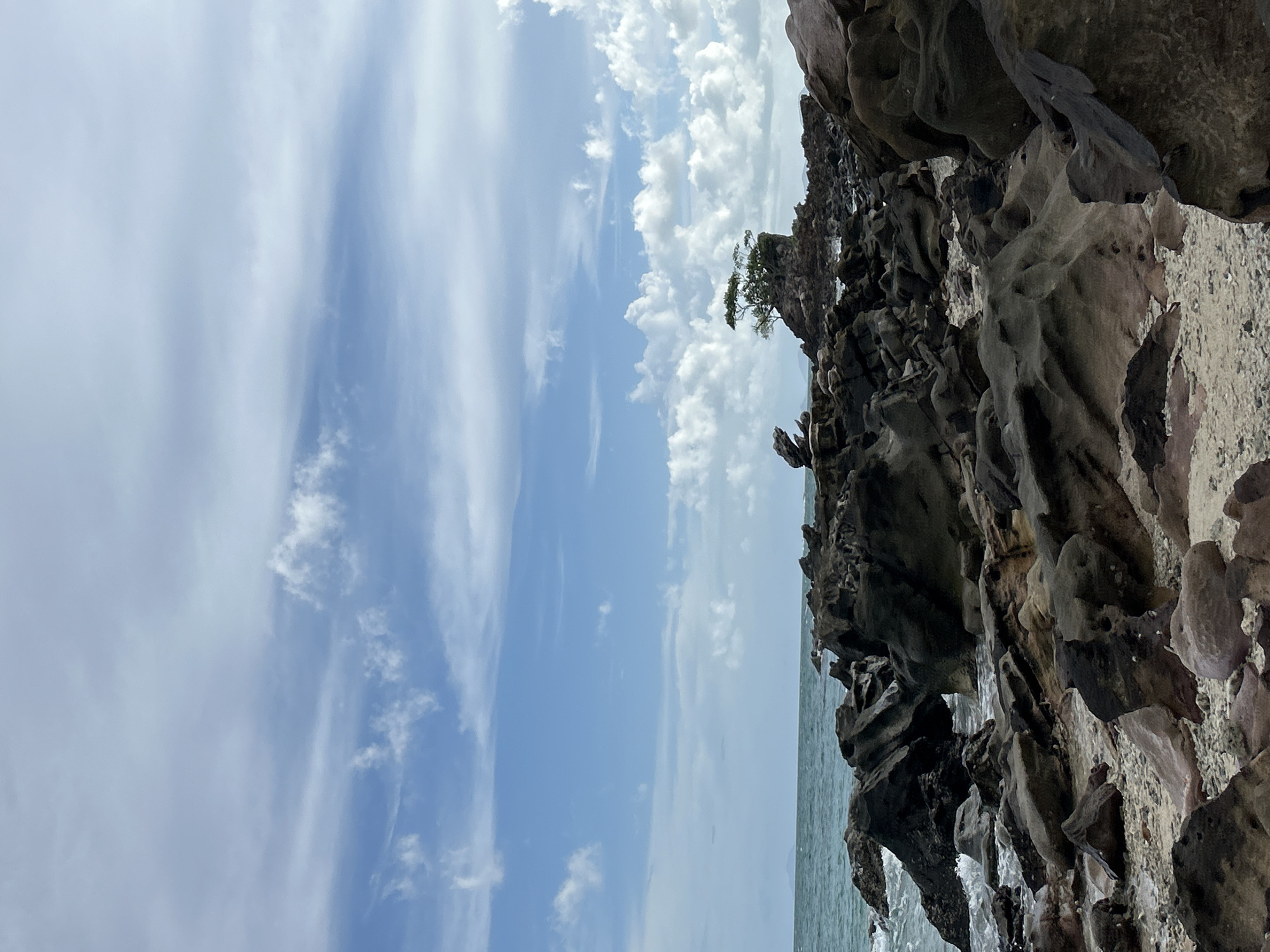 A wide shot captures a rocky shoreline with the ocean and a cloudy sky in Phru Nai, Thailand. The foreground is dominated by dark, weathered rocks of various sizes, some smooth and rounded, others sharp and jagged. Small pebbles and sand are visible between the rocks. In the middle ground, the turquoise ocean meets the shore, with gentle waves lapping against the rocks. A small, rocky islet with a cluster of green trees stands in the water, a short distance from the shore. In the background, the horizon is marked by a distant, hazy landmass. The sky is a mixture of pale blue and white, with wispy clouds scattered across it. There are no people or animals visible in the image.