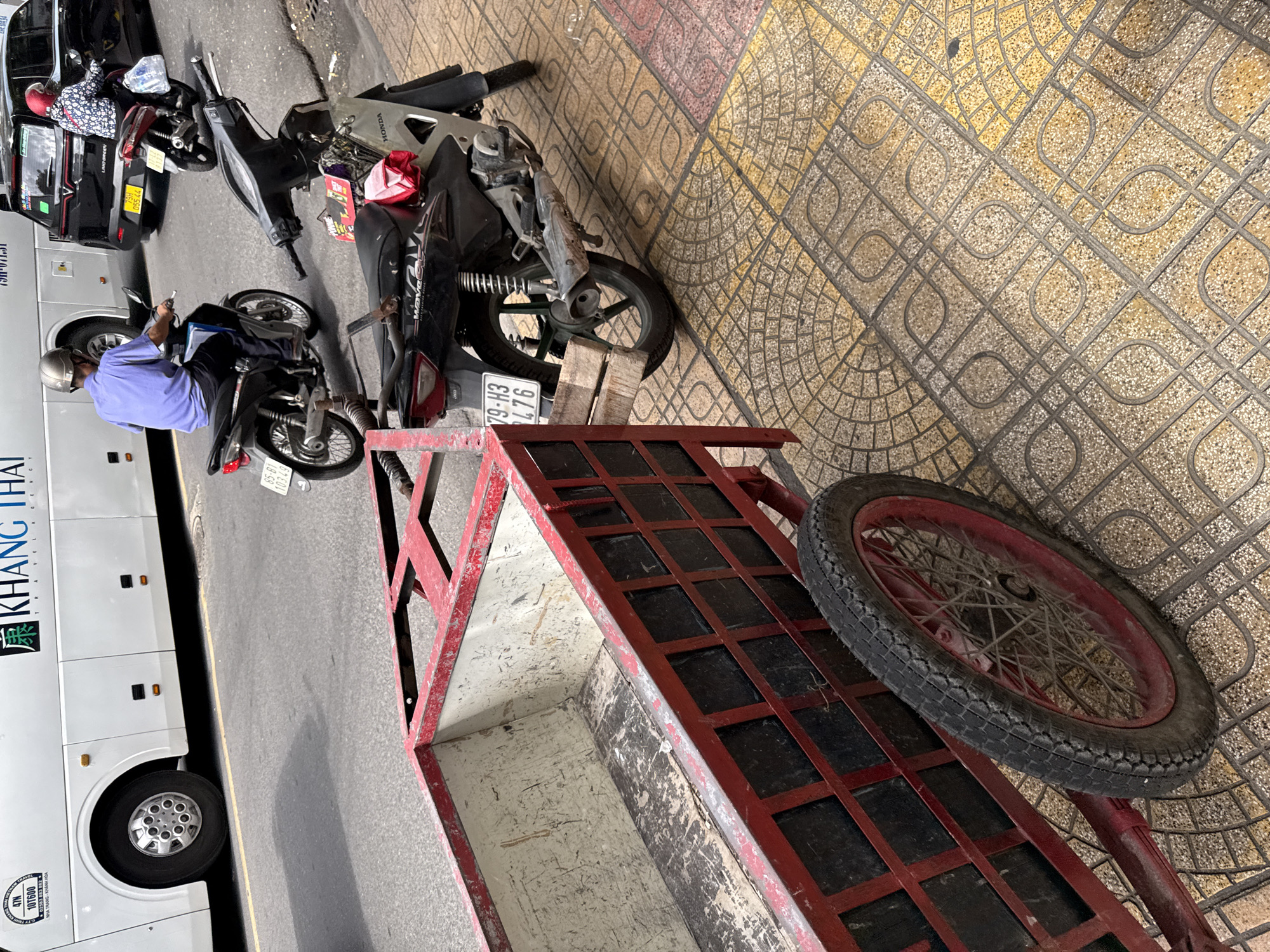 In Nha Trang, Vietnam, a red metal cart with a wooden interior is parked on a sidewalk. The cart has a single spoked wheel with a rubber tire. Behind the cart, a black Honda Wave motorcycle with a license plate reading "79-H3 476" is parked. Further back, a man wearing a blue shirt and helmet is riding a motorcycle. To the right of the man, a black car is visible, and a woman in a red helmet is riding a red motorcycle. In the background, a white bus with "KHANG THAI TRAVEL AGENCY" written on it is parked. The sidewalk is paved with textured yellow and brown tiles.