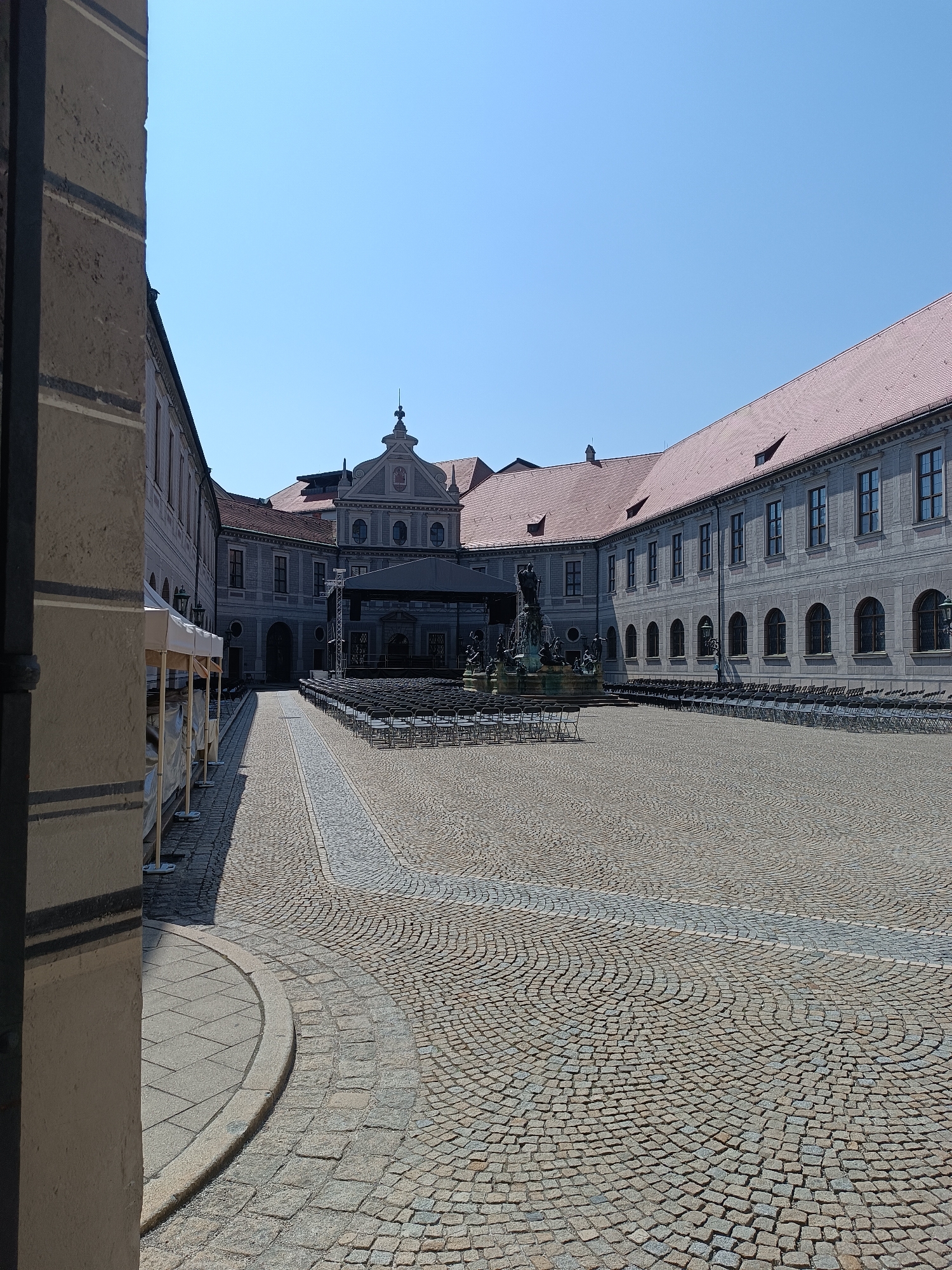 This image depicts a paved courtyard in Munich, Germany, likely a public space or part of a historical complex. The courtyard is laid with cobblestones arranged in a curved pattern. On the right side of the image, a large, ornate building with a red-tiled roof and numerous arched windows spans a significant portion of the frame. Rows of dark chairs are neatly arranged in front of the building, suggesting preparation for an event or performance. In the center of the courtyard, a stage structure with a dark canopy is visible, further indicating an outdoor event. A fountain with sculpted figures stands near the stage. Tents, possibly for vendors or an information booth, are set up on the left side of the courtyard, near a section of a stone building. The sky is clear and blue. The overall scene suggests a sunny day in a historical urban setting, likely prepared for an outdoor gathering.
