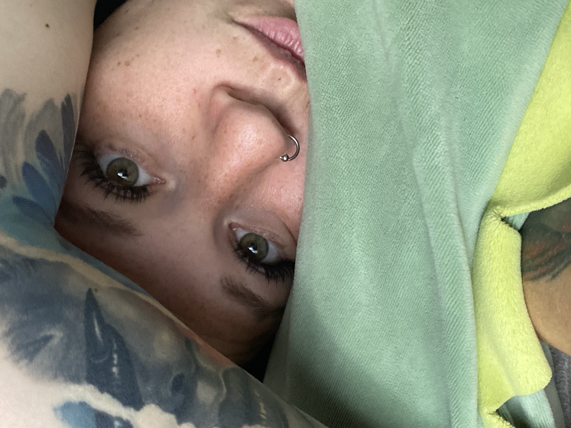 A close-up, overhead shot shows a person's face peeking out from under a light green blanket. The person has light skin with freckles, green eyes with long eyelashes, and a nose ring. A tattoo is visible on their arm, which is partially covered by the blanket. The background is out of focus. This image was not taken in Chaikovsky, Russia.