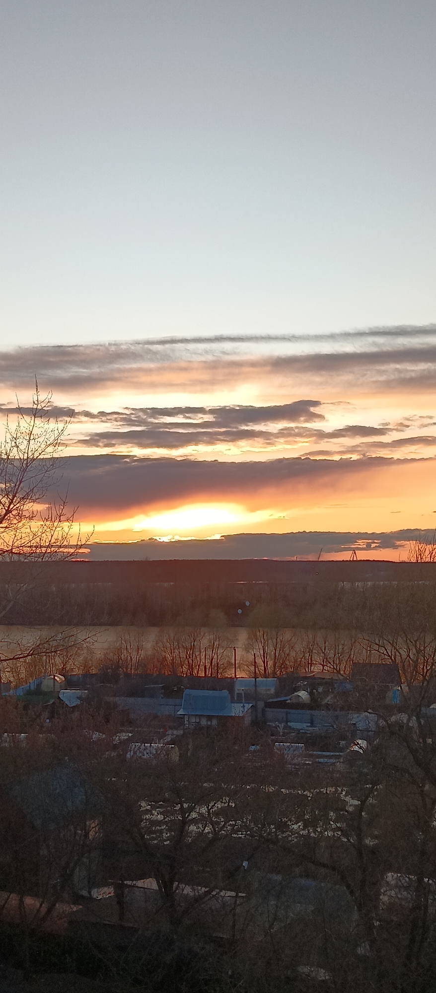 The image presents an expansive landscape view at sunset, looking out over a river and a small settlement. The sky is a prominent feature, transitioning from a light grey-blue at the top to vibrant shades of orange, yellow, and pink near the horizon. Numerous clouds are scattered across the sky, some appearing as long, horizontal streaks, while others are darker and more amorphous, reflecting the warm glow of the setting sun. The sun itself is a brilliant, golden light source positioned low in the sky, just above the distant horizon.
Below the illuminated sky, a wide river stretches across the midground, its surface appearing calm and reflecting subtle hints of the sky's colors. A dense line of bare, deciduous trees borders the river on the opposite bank, creating a dark silhouette against the sunset. Beyond the river and trees, a dark, undulating landmass or forest extends to the far horizon. Faint, dark industrial structures, possibly cranes or towers, are visible in the far distance on the right side of the horizon line.
In the foreground, a collection of small houses or buildings with various roof colors (some blue, some darker) are nestled among bare trees and fences. The intricate network of dark, leafless branches of trees frames the bottom and left sides of the image, partially obscuring the settlement. The lack of foliage on the trees strongly suggests a late autumn, winter, or early spring season. No human or animal activity is visible, contributing to a serene and still atmosphere. There is no discernible text within the image.