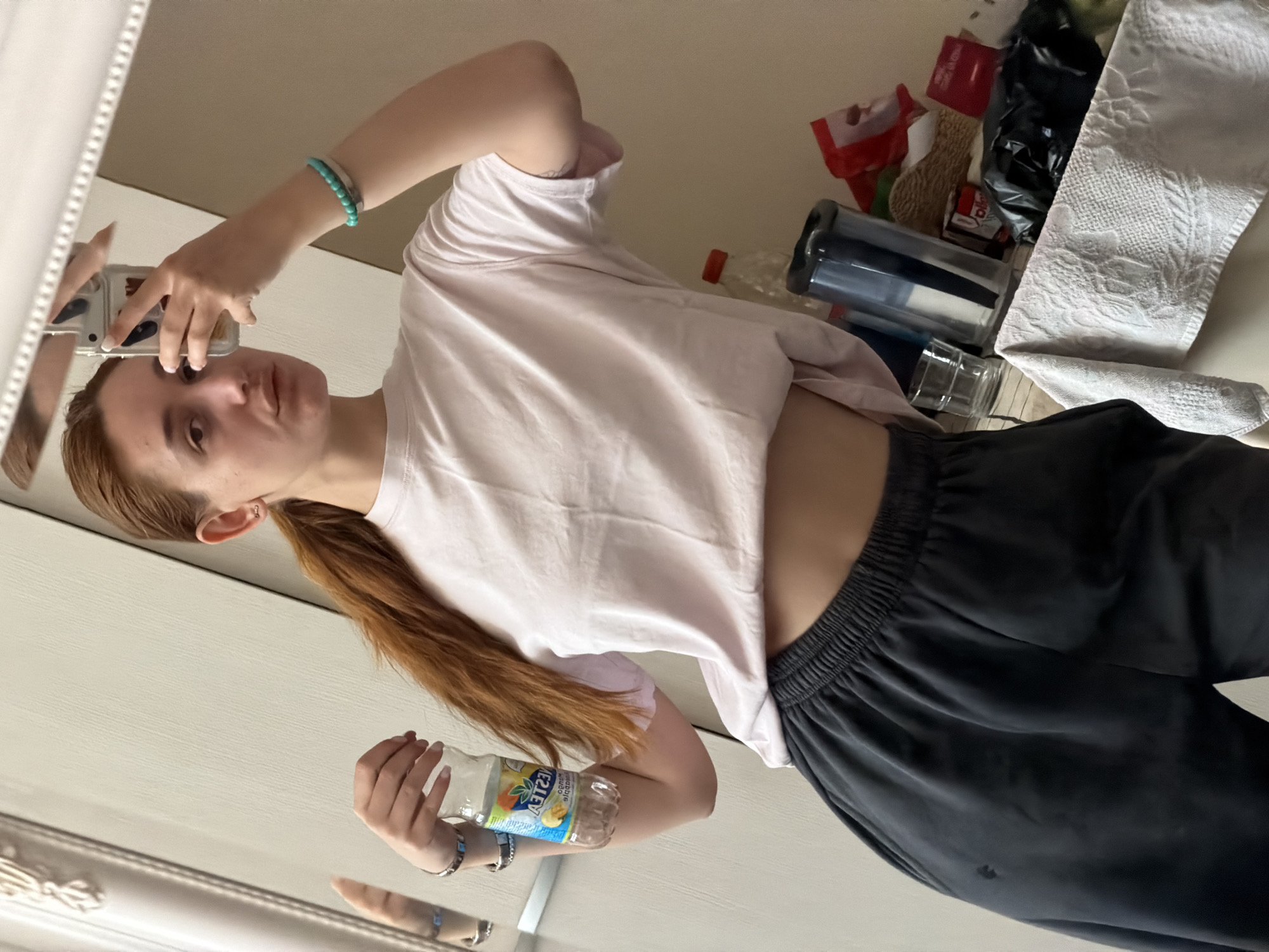 A young woman is shown in a mirror selfie, with the image rotated 90 degrees clockwise. She has fair skin, long reddish-brown hair tied in a low ponytail, and a neutral expression, looking directly at the camera. She wears a light pink, short-sleeved crop top that exposes her midriff, paired with dark grey or black sweatpants. Her left wrist displays a turquoise beaded bracelet and a silver-colored bracelet, while her right wrist has a darker beaded bracelet and another silver-colored bracelet. A small stud earring is visible in her right ear.
She holds a smartphone in her left hand to capture the reflection. In her right hand, she holds a clear plastic bottle with a colorful label. The text on the label is mirrored, but "NESTEA" and "LEMON" are discernible, along with illustrations of pineapple and other citrus fruits.
The setting is indoors, likely a casual home environment in Sofia, Bulgaria. The white, ornate frame of the mirror is visible on the left edge. The background reflected behind the woman is somewhat cluttered, showing various household items on a light-colored surface. These items include a dark tumbler with a clear lid, another clear plastic bottle with an orange cap, a black plastic bag, red packaging, a white patterned towel or cloth, and other indistinct objects. The overall scene depicts a casual, everyday moment.