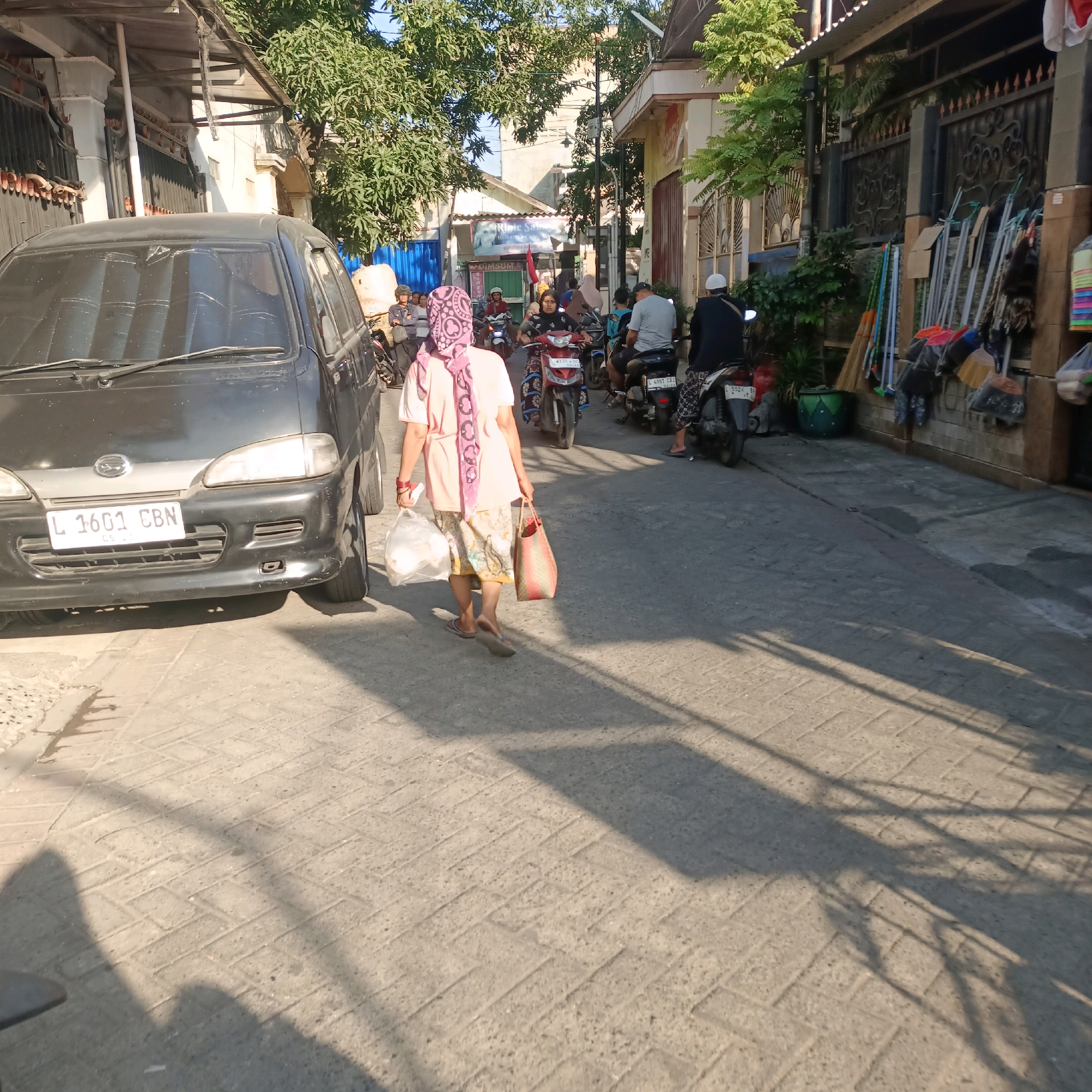 A woman in a pink headscarf and a pink shirt walks away from the camera, carrying two bags. She is on a paved street in Surabaya, Indonesia, with a dark-colored van on her left and a line of motorcycles on her right. Buildings with signs and trees line the street. The scene is lit by sunlight, casting long shadows on the ground.