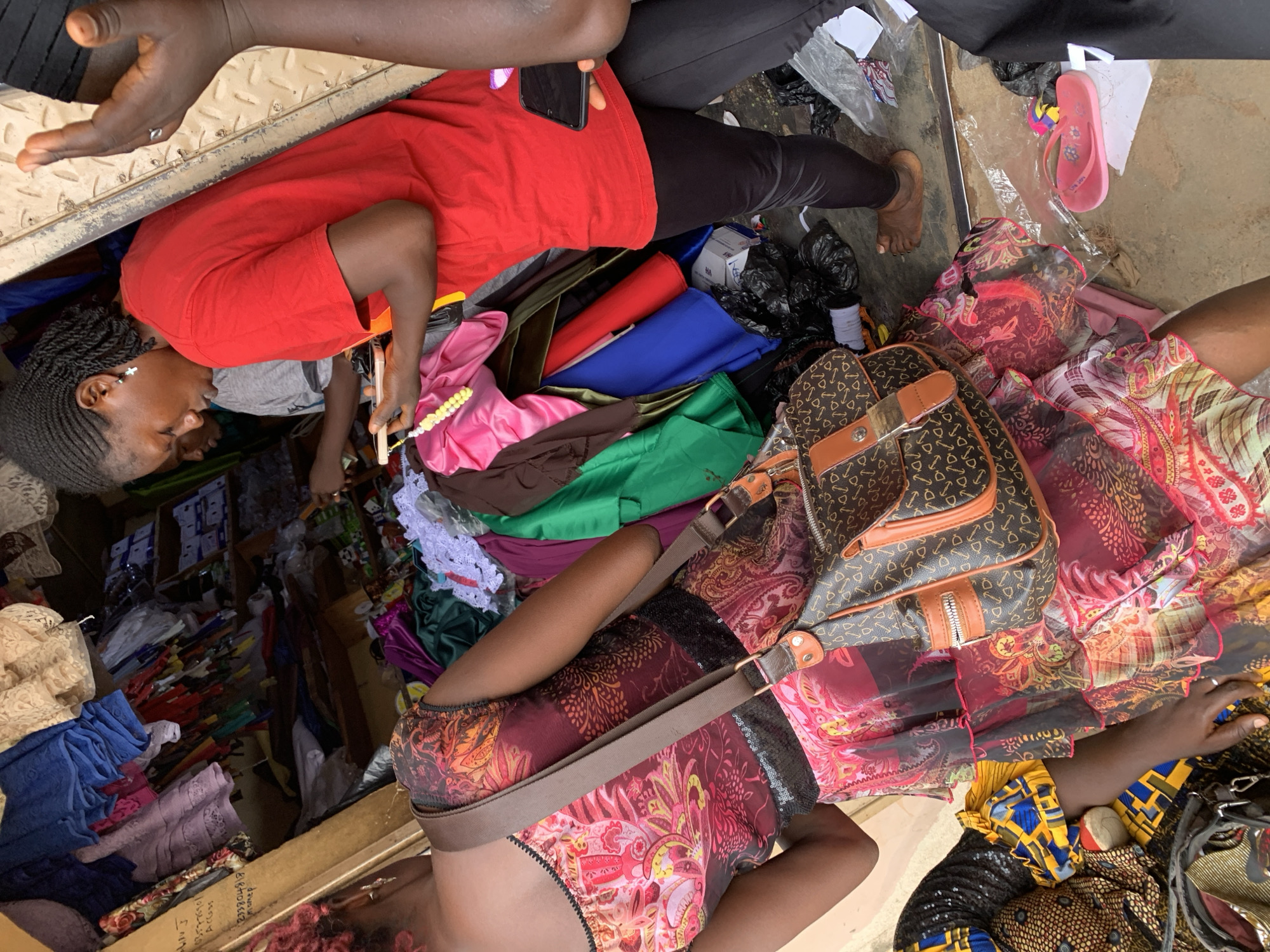 The image shows an open-air market scene. Several women are present, dressed in a variety of clothing, examining fabrics and clothing items. The fabrics and items vary in color and pattern and are stacked and spread across the space. A woman in a red shirt holds a beaded string and interacts with another woman seated near rolls of fabric. A handbag sits on top of the fabric rolls. A pink flip-flop is visible on the ground amidst packaging and trash. The setting appears to be in a busy market location.
