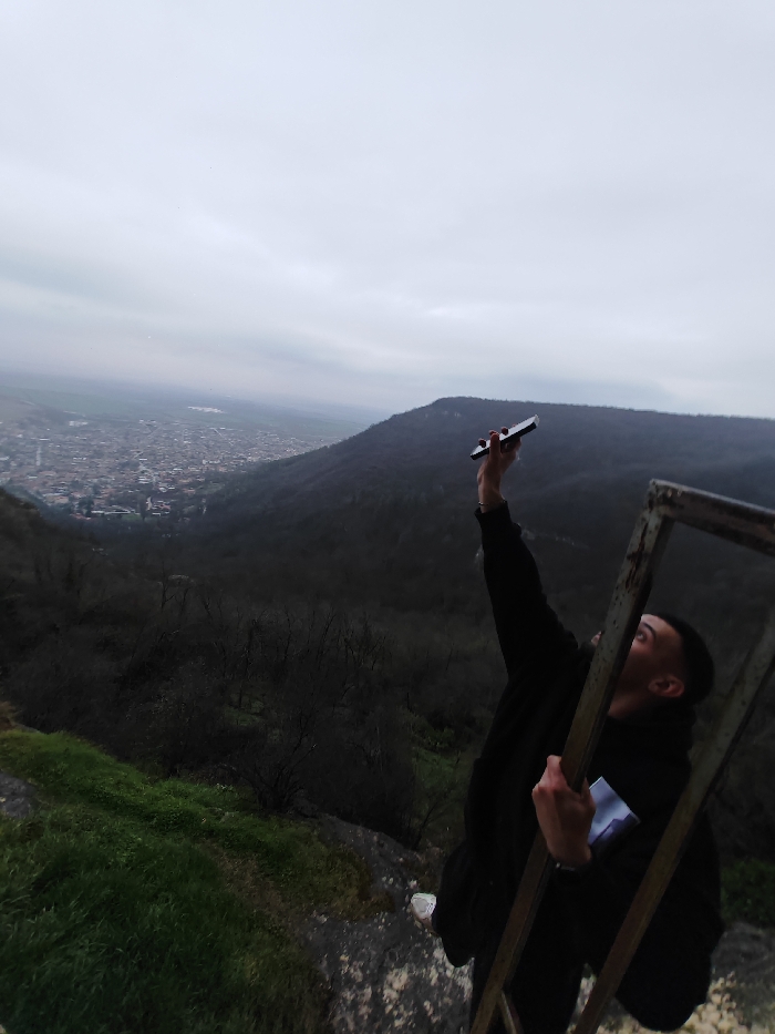 The image shows a young male on a rocky, partially grass-covered elevated position overlooking the city of Shumen, Bulgaria, under an overcast sky. The male, wearing a dark hooded sweatshirt, extends his right arm vertically, holding a dark smartphone skyward. His left hand grips a vertical bar of a rusty, dark metal frame situated prominently on the right side of the frame. His head is tilted back, and his mouth is open, with his gaze directed upwards. Below the immediate foreground, dark, densely forested hillsides descend into a valley where the urban sprawl of Shumen is visible, characterized by numerous buildings. Further in the distance, fields and a faint body of water are discernible under the uniformly gray, cloudy sky. The scene is consistent with a viewpoint from the Shumen Plateau.