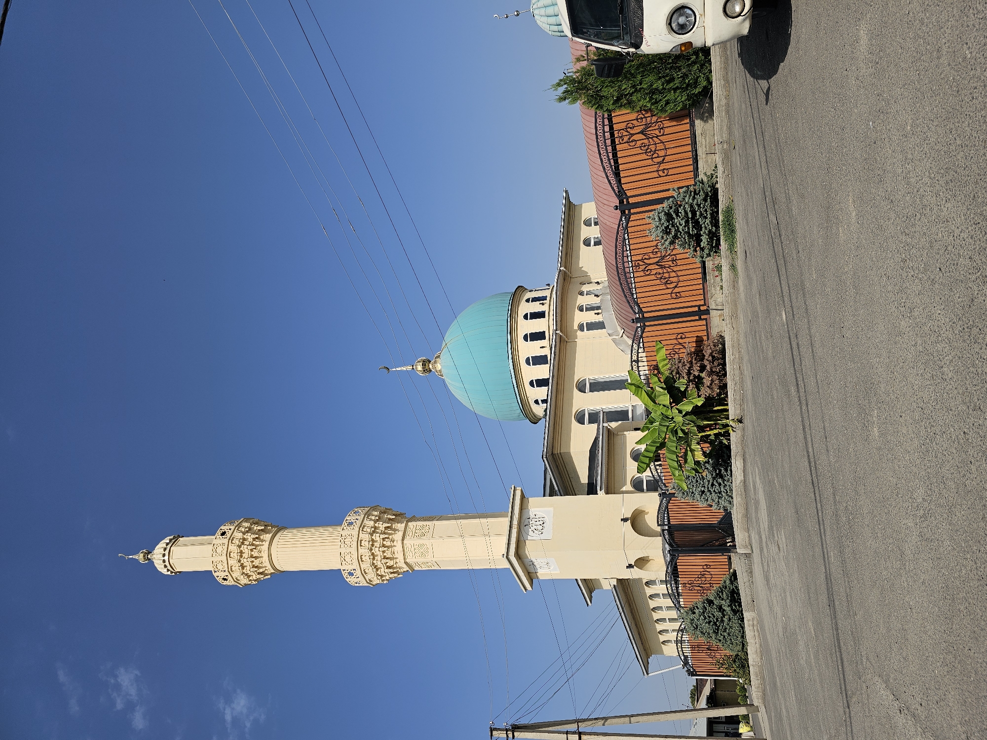 The image shows a mosque in the city of Özgön, Kyrgyzstan. The mosque features a tall, cream-colored minaret with intricate decorative elements and a crescent moon at its apex. Adjacent to the minaret is the main building of the mosque, characterized by its light yellow facade, arched windows, and a prominent turquoise dome topped with a golden finial. A decorative fence, predominantly orange with black wrought iron accents, encloses the mosque's grounds. Several small evergreen trees and banana plants are visible within the fenced area. A white van is parked on the right side of the image, partially obscuring a smaller dome structure and vegetation. The foreground consists of an asphalt road, with utility poles and overhead power lines visible against a clear blue sky. The scene is captured during daylight with no people present.