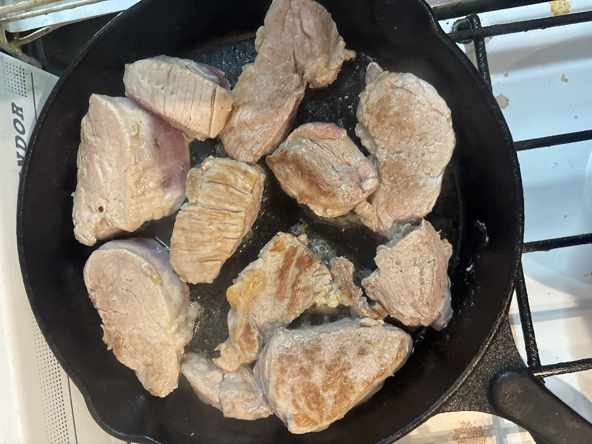 The image shows several pieces of meat cooking in a black cast iron skillet on top of a white gas stove. The meat appears to be pork tenderloin, cut into thick medallions and seared. The skillet is sitting on a burner, surrounded by parts of the stove's grates. The location is difficult to determine from the image alone, and impossible to relate to the city of Buenos Aires, Argentina, without additional information.