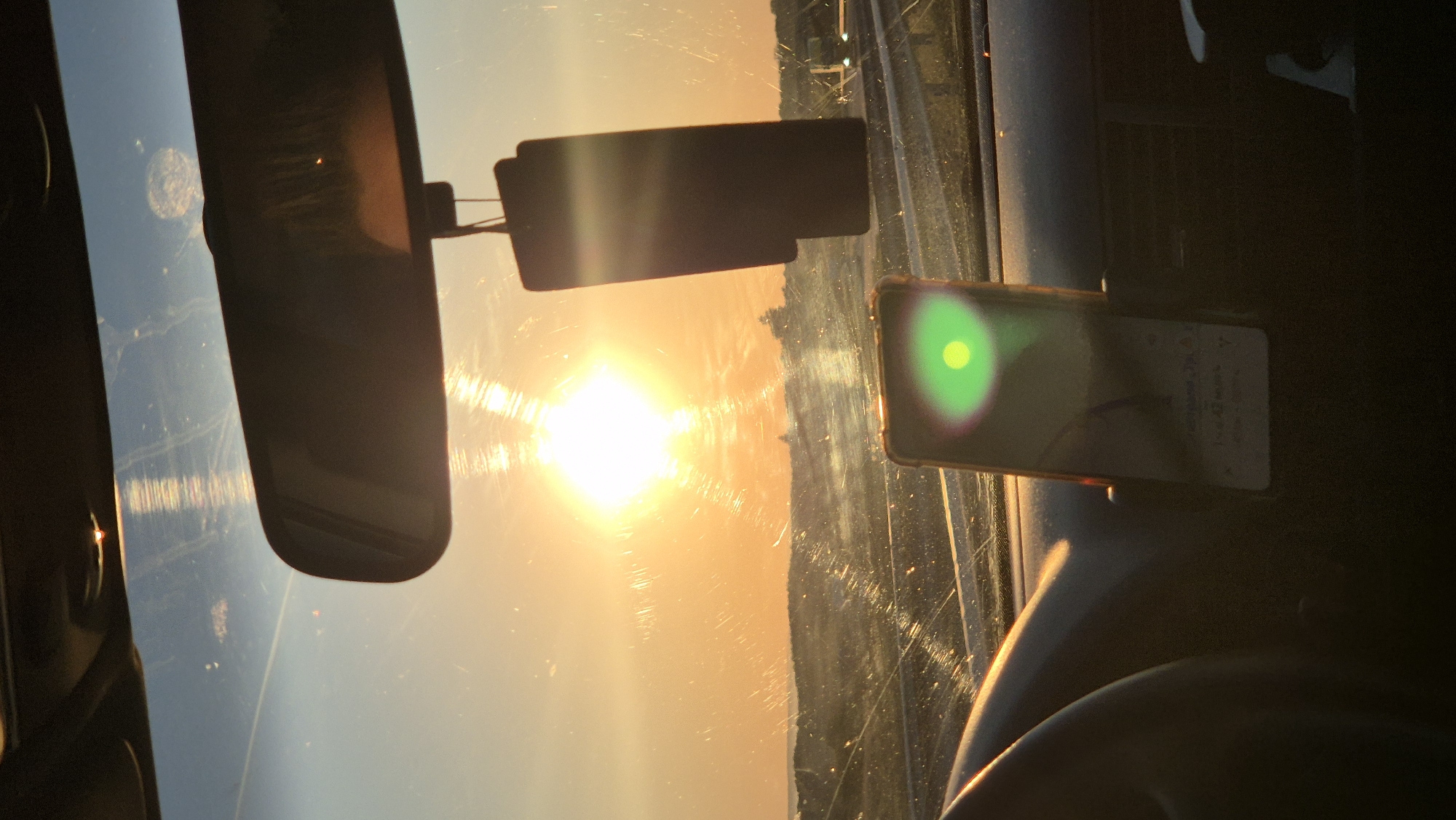 The image captures a sunset viewed from inside a vehicle. The sun is a bright, intense orb in the sky, casting a warm, orange glow across the scene. The view is through the windshield, which shows some streaks and dust. A rearview mirror is visible at the top center of the frame, and a dark, rectangular object, possibly an air freshener, hangs from it. In the foreground, a portion of the car's dashboard and steering wheel is visible on the left. A smartphone, mounted on a holder, is positioned on the dashboard in front of the passenger seat, displaying a navigation interface with a green light flare reflected on its screen. In the distance, a landscape with some vegetation and possibly some structures can be seen under the dramatic sunset sky. The image does not contain enough visual information to determine the specific location within the city of Kabile, Bulgaria.