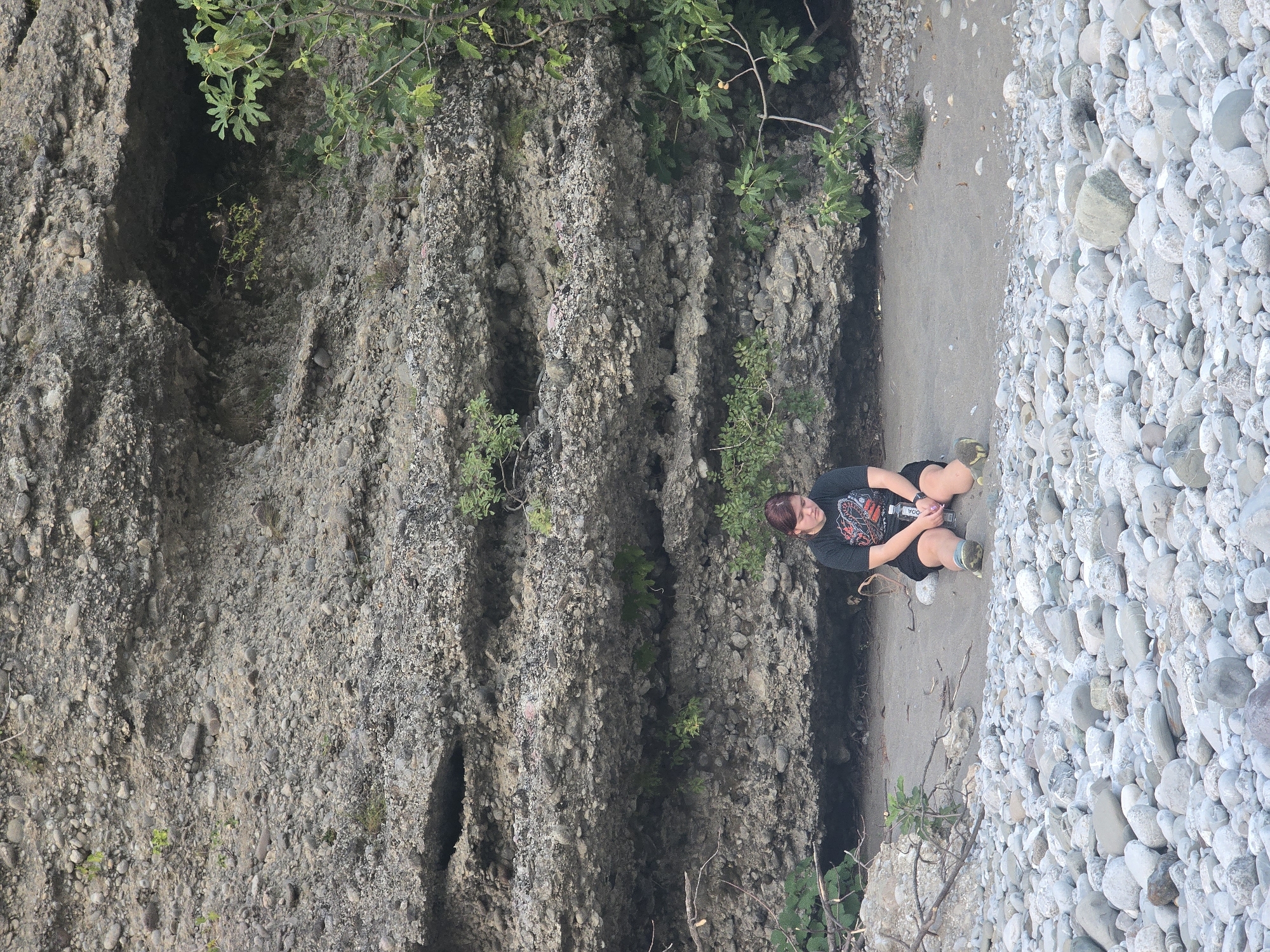 A young woman with reddish-brown hair is sitting cross-legged on a sandy patch of ground at the base of a large, layered rock formation. She is wearing a dark t-shirt with a graphic print and dark shorts. Her arms are resting on her knees, and she is holding a clear plastic water bottle. Her gaze is directed downwards. The rock formation behind her is textured with various sizes of embedded stones. Small green plants are growing in crevices and at the base of the rock. The ground in the foreground is covered with numerous smooth, light-colored stones. To the right, a branch of a leafy green tree extends into the frame, bearing small green fruits. The scene appears to be outdoors, likely near a body of water, given the prevalence of stones. The overall environment suggests a natural, somewhat rugged setting in Bioče, Montenegro.