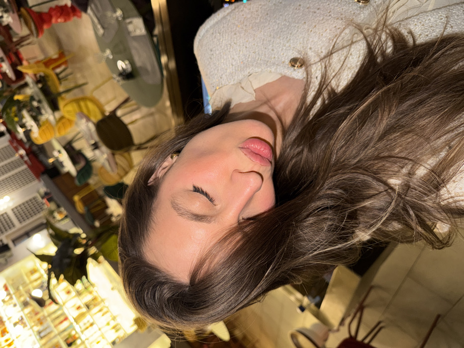 A woman with light skin and closed eyes is the main subject. Her dark brown hair with some lighter streaks cascades across a tweed jacket with gold buttons. Her lips are full and her face has a subtle, rosy tint. The background shows blurred interior elements, including tables, chairs, plants, and a display case full of various items, suggesting a store or restaurant setting. The image is rotated.