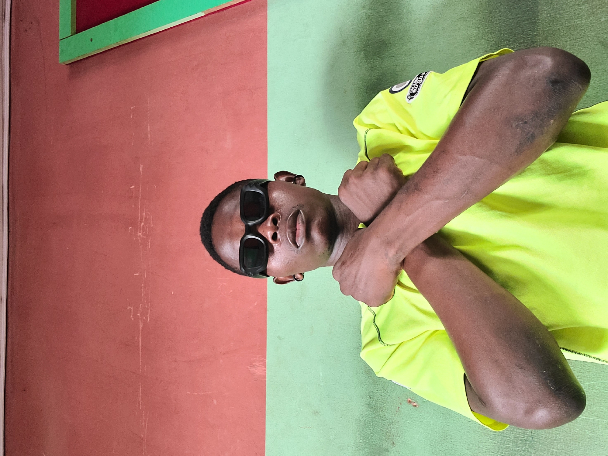 A young Black man wearing black sunglasses and a neon yellow t-shirt stands with his arms crossed over his chest in front of a wall divided horizontally into a reddish-brown upper section and a light green lower section. A sliver of a red rectangle outlined in green is visible in the upper right corner of the frame. The man is looking directly at the camera with a neutral expression. His left arm is over his right arm. The wall appears to be outdoors, possibly a building exterior in Gura Top, Nigeria.