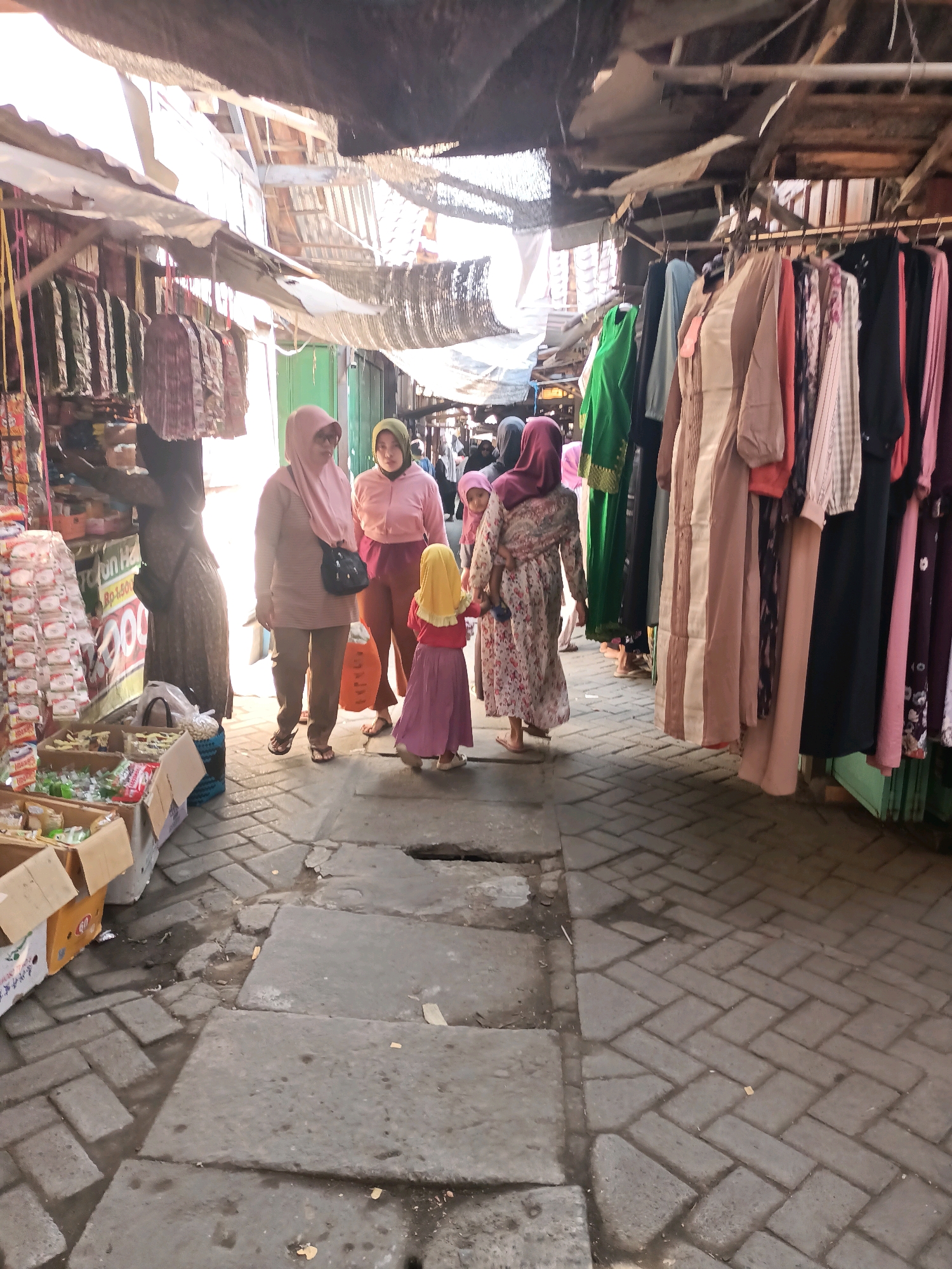 This is a forensic, matter-of-fact description of an outdoor market scene in Badas, Indonesia. **Location:** Outdoor market stall area, Badas, Indonesia. **Environment:** The scene depicts a narrow, outdoor walkway lined with market stalls. The ground is paved with interlocking bricks and some larger rectangular concrete slabs, with visible patches of dampness and debris. Overhead, a makeshift roof structure made of corrugated metal and tarps provides partial shade. The stalls are open-fronted, displaying merchandise. One stall on the left sells packaged goods stacked in boxes and on shelves, including what appears to be snacks or small food items. Another stall on the right displays a variety of clothing, mostly dresses or long garments, hanging from racks. The overall atmosphere suggests a busy, functional marketplace. **People:** There are at least five individuals visible, all appearing to be women or girls, and all wearing hijabs or headscarves. * On the far left, a woman in a dark brown patterned dress and headscarf is partially obscured, appearing to be attending to or browsing items at the stall. * A woman in a light pink hijab and a greyish-pink patterned long-sleeved top, wearing brown trousers, walks towards the right of the frame, looking forward. She carries a dark bag over her shoulder. * Next to her, a woman in a light green hijab and a light pink long-sleeved top, wearing darker trousers, also walks towards the right, looking forward. * In front of these two women, a young girl wearing a bright yellow hijab and a pink dress walks ahead, her back to the camera. * Further to the right, partially visible behind other individuals, is a woman in a floral-patterned dress and a pink hijab, carrying a baby or young child on her left side. The child is wearing a pink outfit and appears to be looking towards the left. **Objects and Actions:** * The market stalls are stocked with goods. The stall on the left has rows of packaged items, with a large price tag or advertisement visible. Boxes of products are on the ground in front of this stall. * The stall on the right features a rack of hanging garments, predominantly in muted and earthy tones, with some brighter colors like green and coral. * The individuals are walking along the market path, suggesting they are shoppers or vendors moving between stalls. The interactions are subtle, with individuals generally moving in