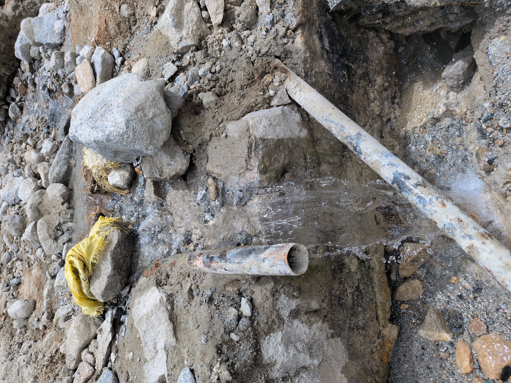 The image depicts an outdoor scene in Panamik, India, characterized by a rocky and muddy terrain. A broken pipe is visible, with water gushing out and flowing downwards. The pipe appears to be made of metal and shows signs of rust and wear. Several rocks of varying sizes are scattered around the pipe and the surrounding area. A yellow cloth is placed on top of a rock near the source of the water flow. The overall environment suggests a site where some sort of construction or repair work might be taking place, or a natural occurrence involving underground water sources. The lighting suggests it is daytime.
