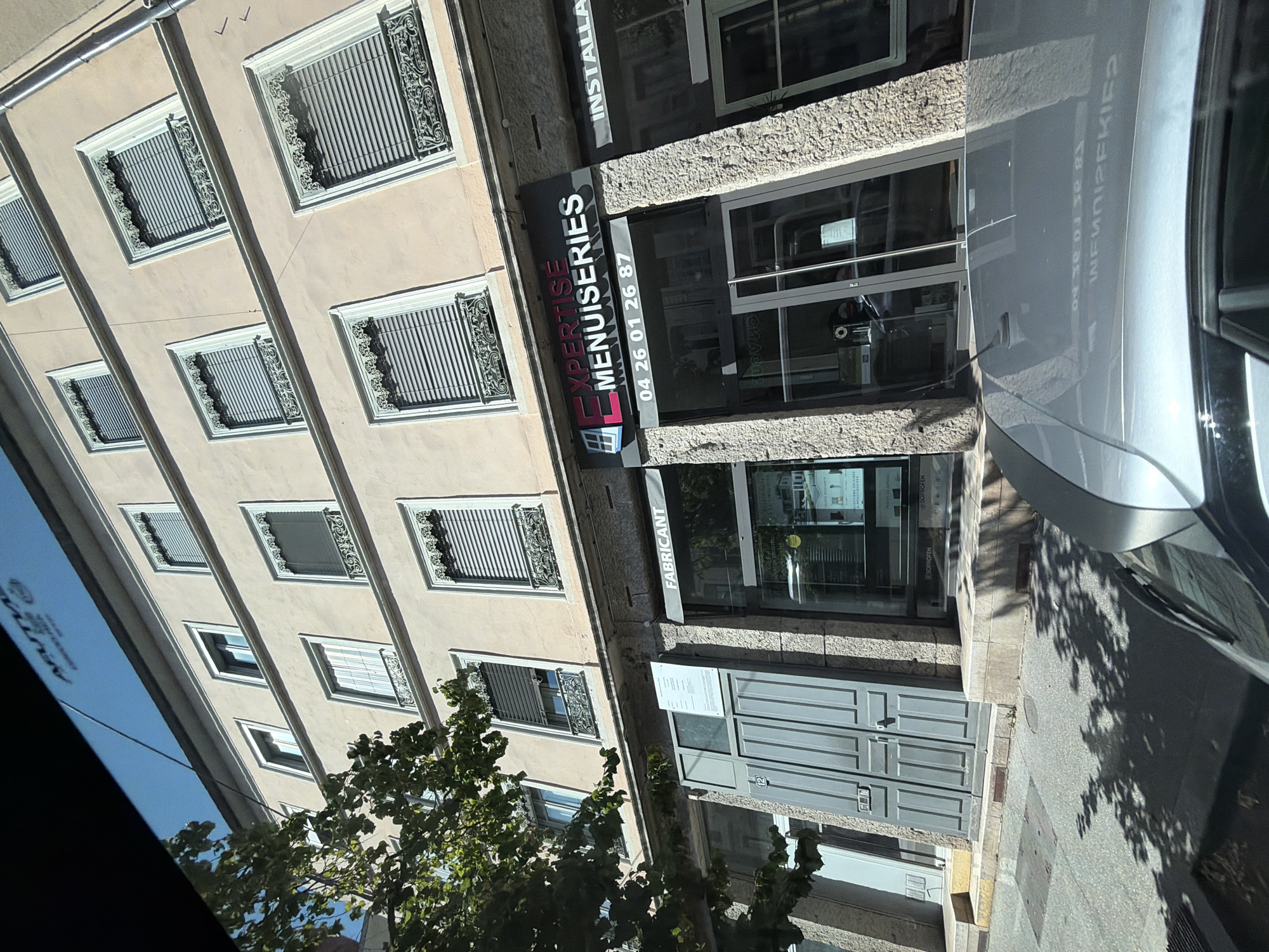 This is a photograph taken in Lyon, France, likely during the daytime under clear weather conditions, as indicated by the bright sunlight and sharp shadows. The image captures a street-level view of a building with a pale yellow facade. The building has multiple windows, many of which are covered by light-colored blinds and feature decorative iron grilles.
The main subject in the foreground is a storefront with a stone-textured facade. A dark gray awning stretches across the top of the storefront, bearing the white text "FABRICANT". Below the awning is a large glass door and windows, offering a glimpse into the interior.
To the left of the storefront, a prominent sign is visible, reading "EXPERTISE MENUISERIES" in white letters on a dark background, with a red "E" logo. Beneath this, a phone number "04 26 01 26 87" is also displayed. Adjacent to the storefront, a silvery car is parked, its reflection visible on the building's glass.
The street in front of the building is paved with asphalt and includes sidewalks and grates. Shadows of trees are cast across the pavement, suggesting the presence of greenery nearby. The overall scene depicts a commercial establishment on a city street.