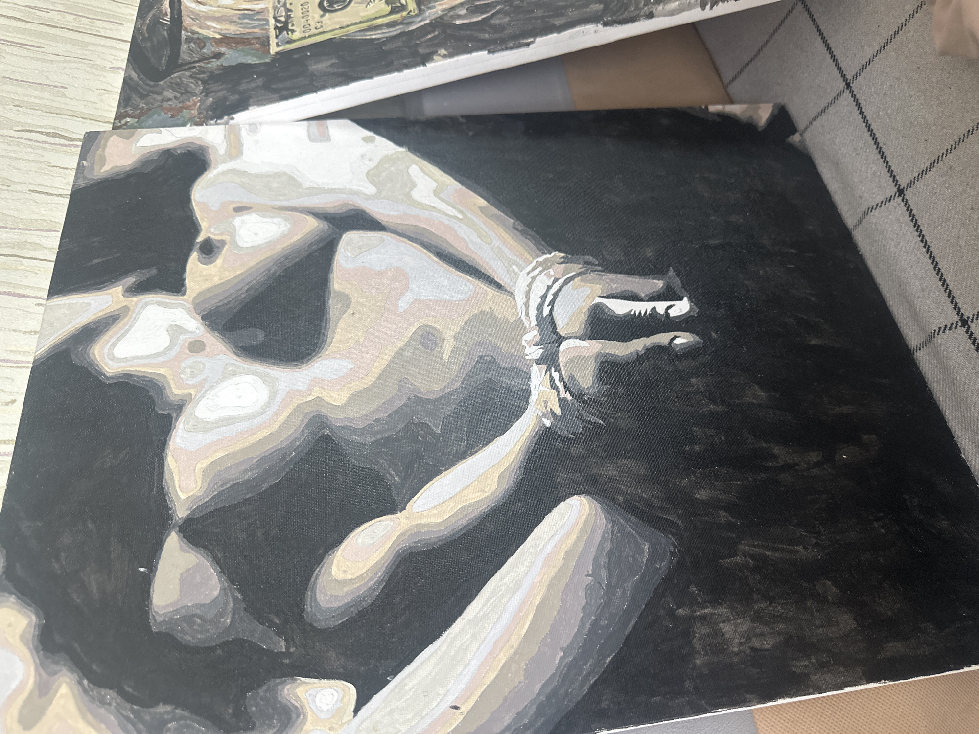 The image shows a painting of a nude figure with their hands bound. The painting uses shades of gray, white, and black to depict the figure against a dark background. Another painting, partially visible in the upper part of the frame, includes an image of a US dollar bill. The location in the city of Vysokiy, Russia is not ascertainable from the image.