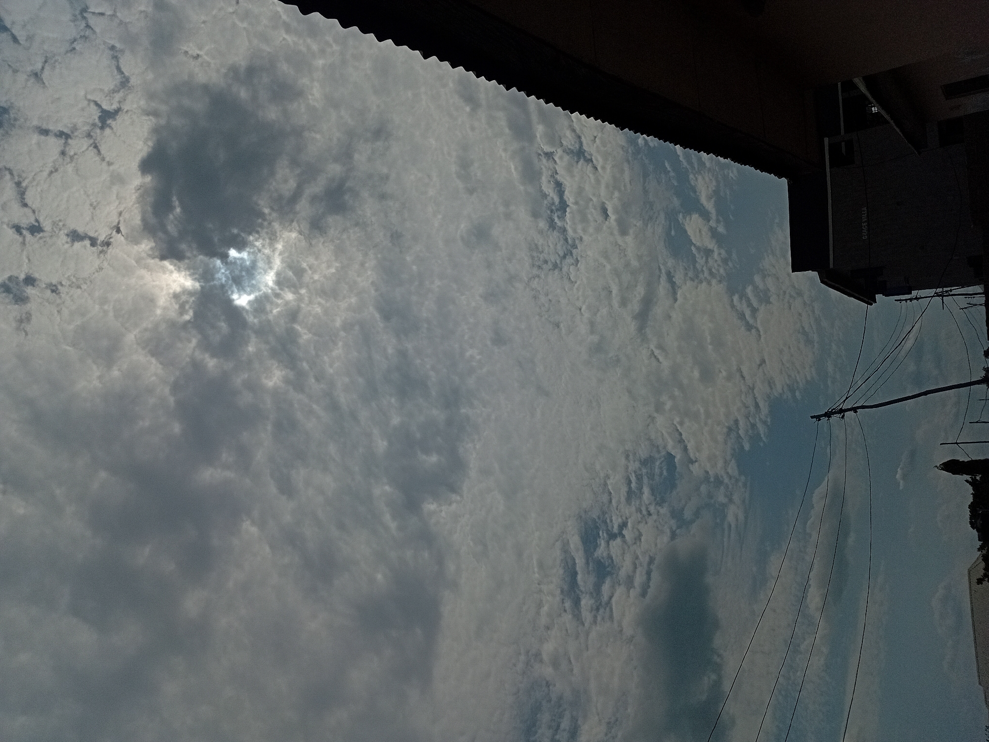 The image, taken from ground level in an urban environment, features a view towards the sky partially obscured by an overhanging roof. The sky is heavily overcast with varying shades of grey and white clouds. There is a hint of sunlight breaking through a denser cloud cover. In the lower right of the image, there are elements of a building, including electrical wires and some vegetation. The location is Unknown, Unknown. No people are visible in the image.